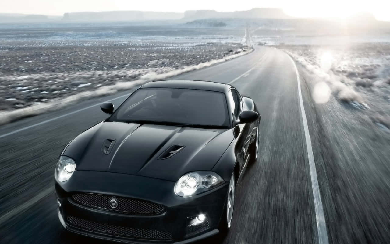 Jaguar Xkr 桌布 壁紙 — 一輛時尚的黑色跑車在陽光明媚的沙漠蜿蜒道路上飛馳，遠處可見平頂山。