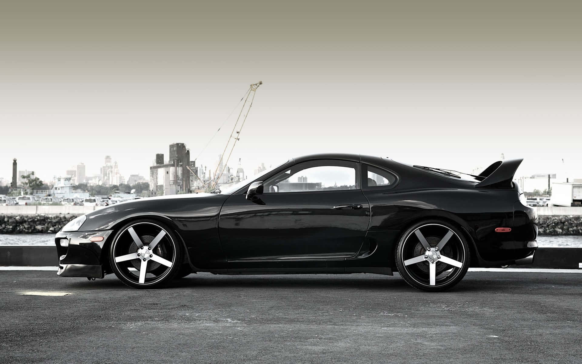 Supra 桌布 壁紙 — 一輛時尚的黑色跑車配銀色輪圈，停在海濱道路上，背景是城市天際線和起重機。