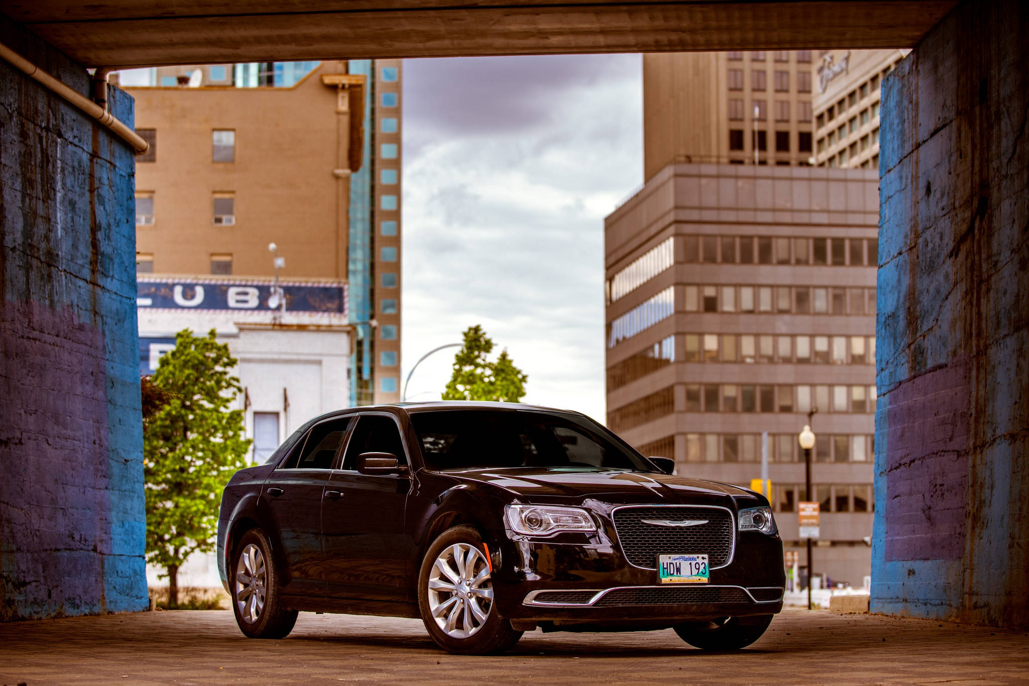 2016 Chrysler 300 壁紙 — 一輛時尚的黑色轎車停泊在藍色城市高架橋下，背景有城市建築物和樹木。