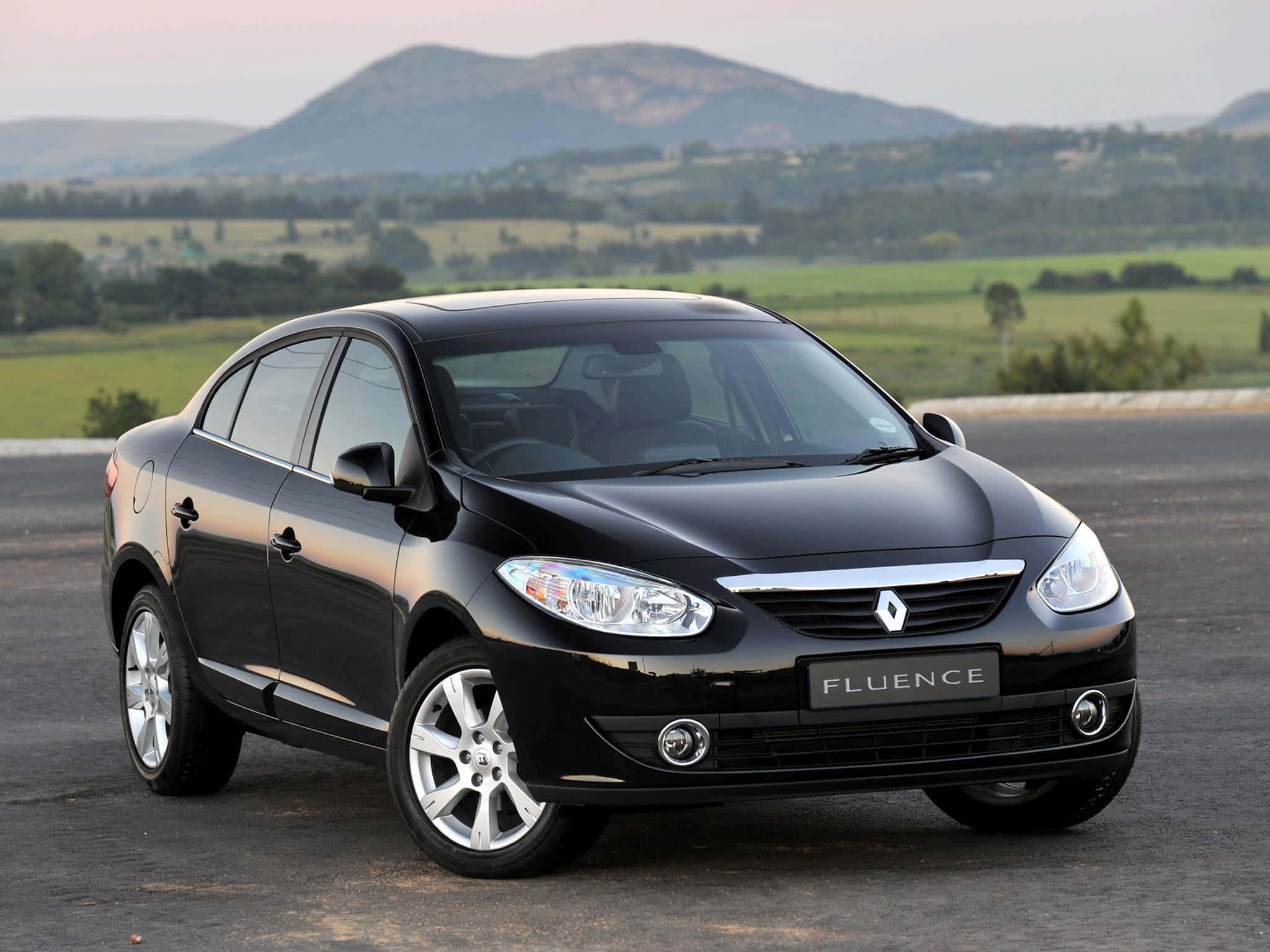 Renault Fluence 桌布 壁紙 — 一輛時尚的黑色雷諾 Fluence 房車停在瀝青路上，背景是起伏的綠色山丘和遠處的山脈，天空呈現柔和的暮色。