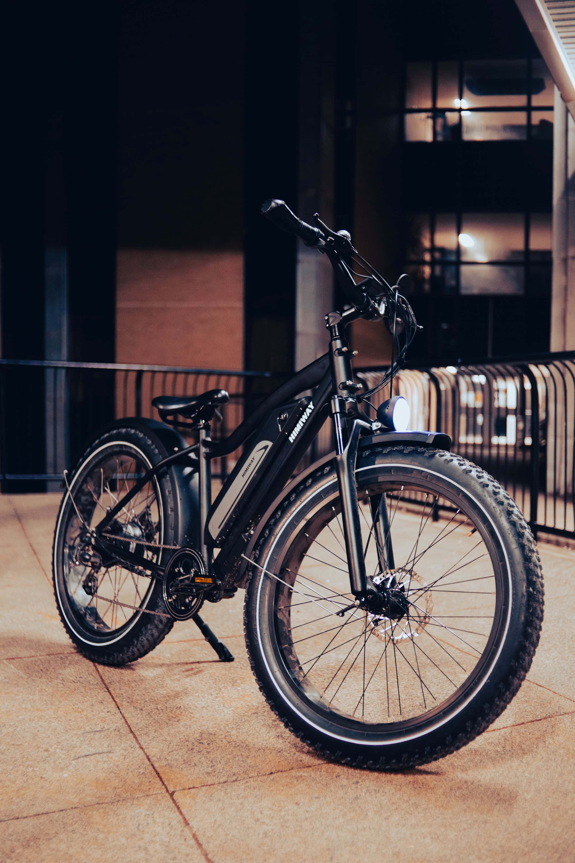 Electric Bike 桌布 壁紙 — 一輛時尚的黑色電動單車配備厚輪胎，夜晚停泊在瓷磚行人路上，附近有一座現代建築物，室內燈光溫暖。