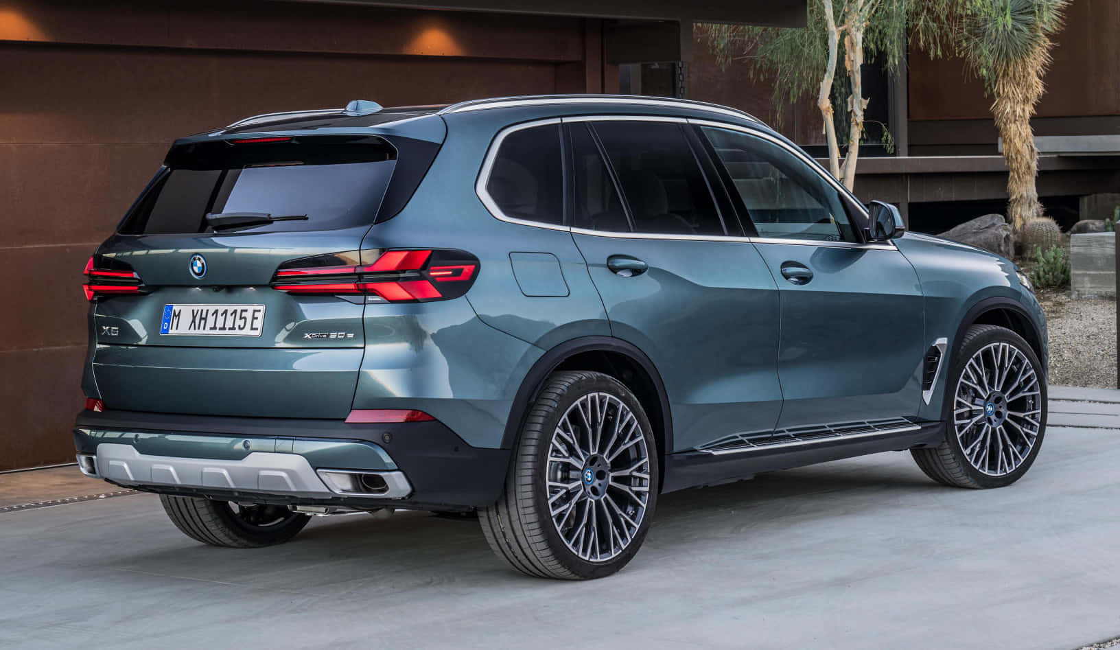 Bmw X5 桌布 壁紙 — 一輛時尚金屬藍色BMW X5 xDrive40e SUV在明亮日光下停放在戶外，背景有現代建築和棕櫚樹。