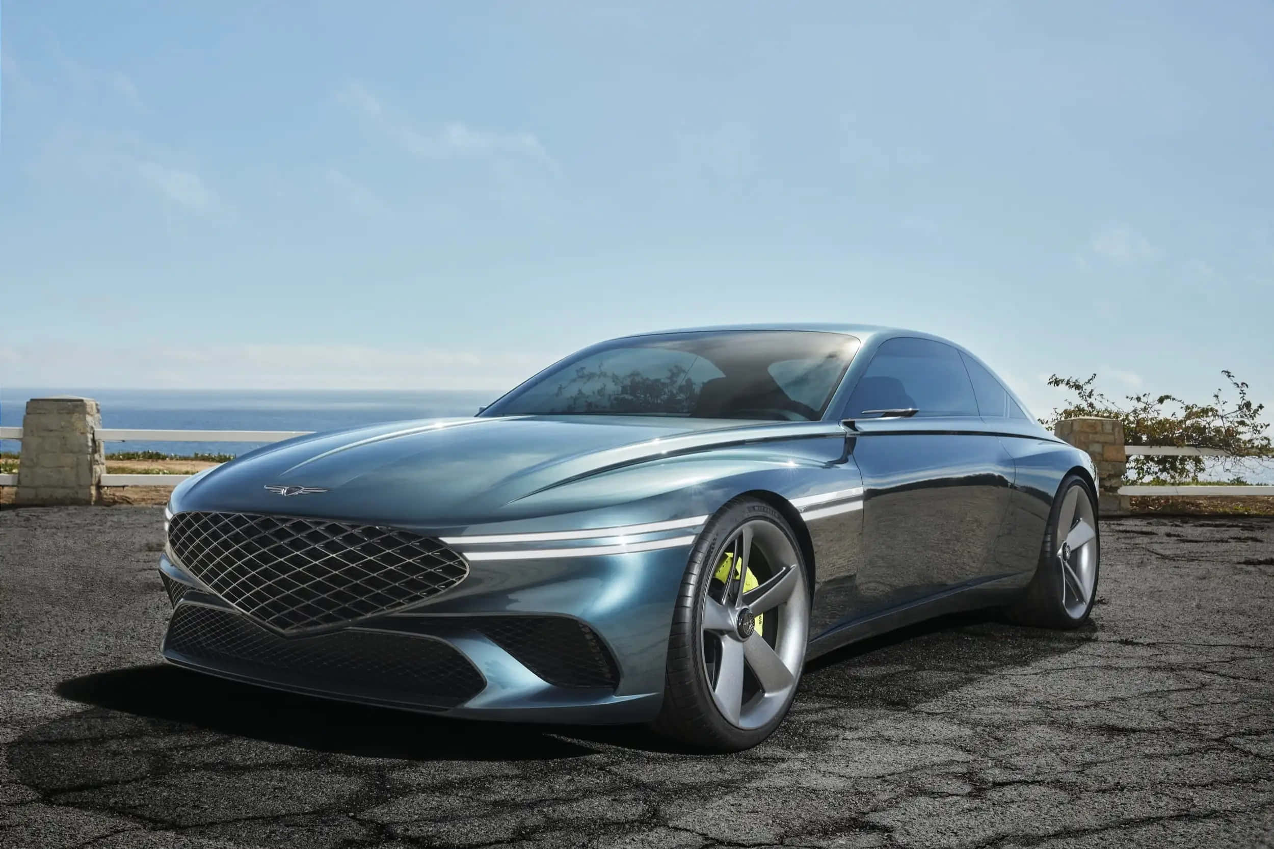 Genesis X Concept 桌布 壁紙 — 一輛時尚金屬藍色跑車配備黃色制動卡鉗，停在裂開的瀝青觀景點，在晴朗天空下欣賞海景。