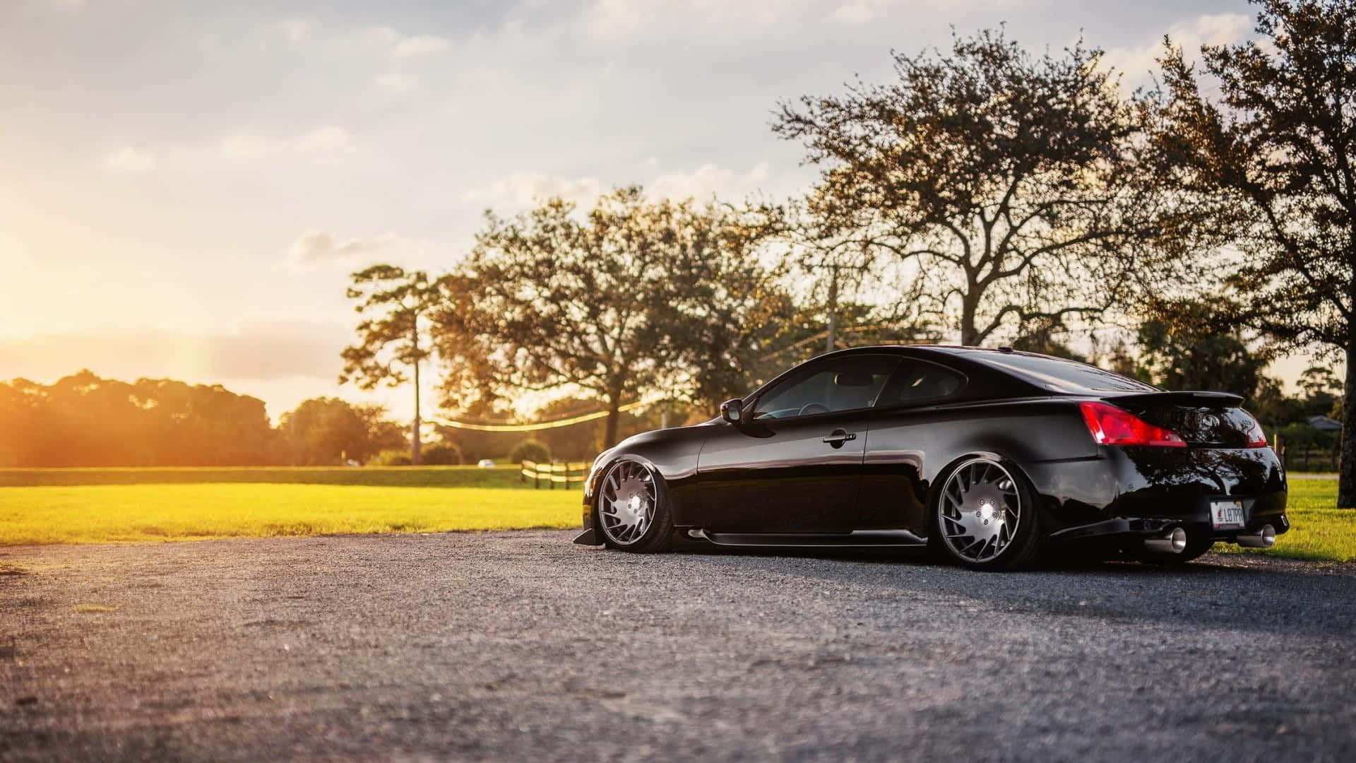 Infiniti G35 桌布 壁紙 — 一輛時尚黑色跑車配備訂製輪圈，在日落時停泊在瀝青路上，在長滿樹木的草地上投下長長的影子。