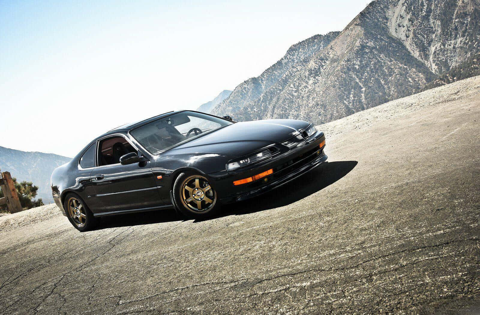 Honda Prelude 桌布 壁紙 — 一輛時尚黑色跑車配金色輪圈，停在龜裂的瀝青路上，背景是崎嶇的山脈和明亮的天空。
