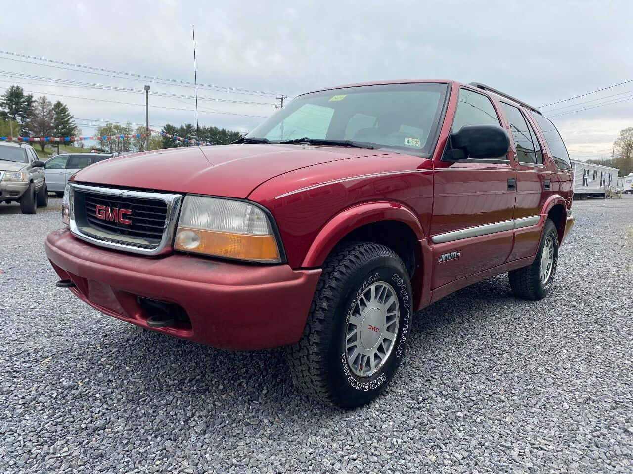 Gmc Jimmy 桌布 壁紙 — 一輛栗色GMC Jimmy SUV停泊在碎石上，背景有樹木和陰天。