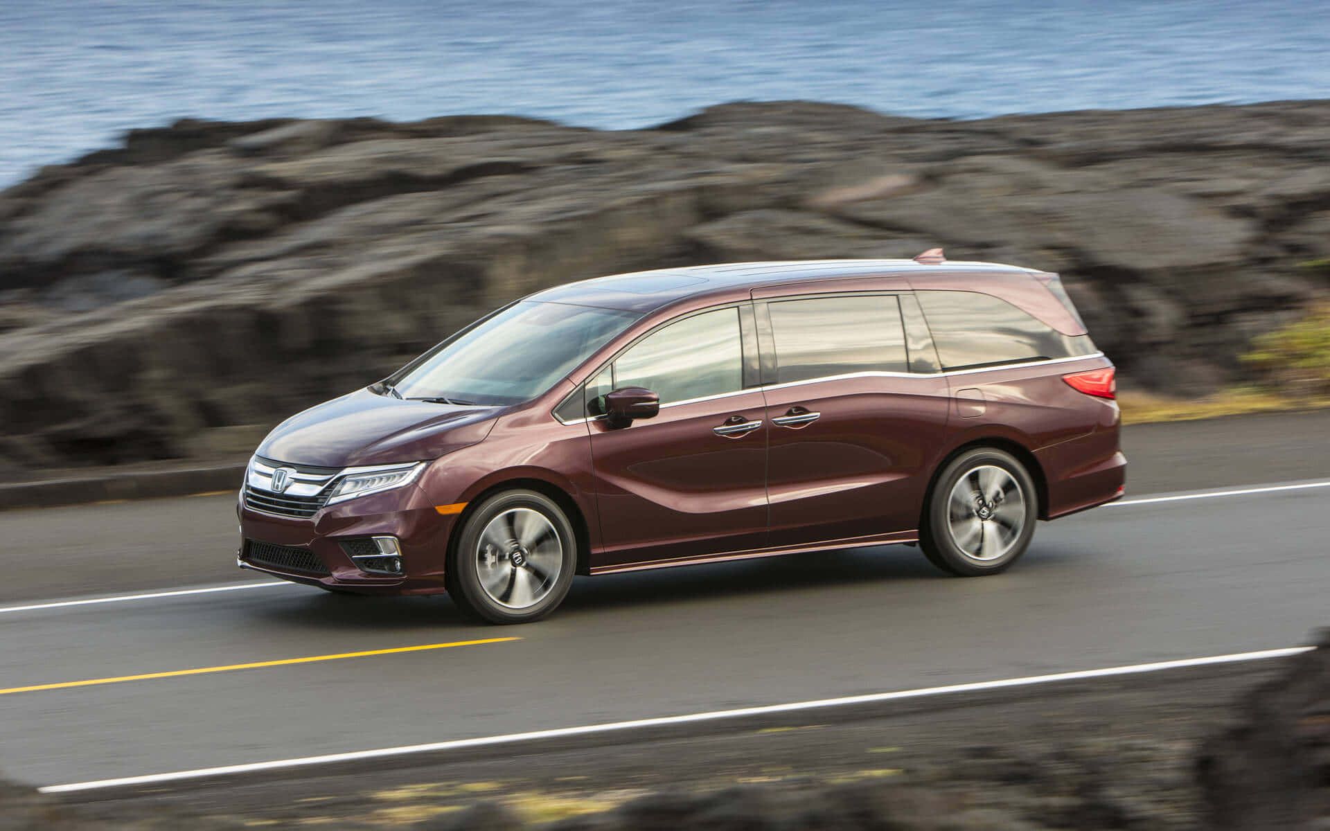 Honda Odyssey 桌布 壁紙 — 一輛栗色小型貨車沿著海岸公路疾馳，背景是模糊的岩石懸崖和海洋。