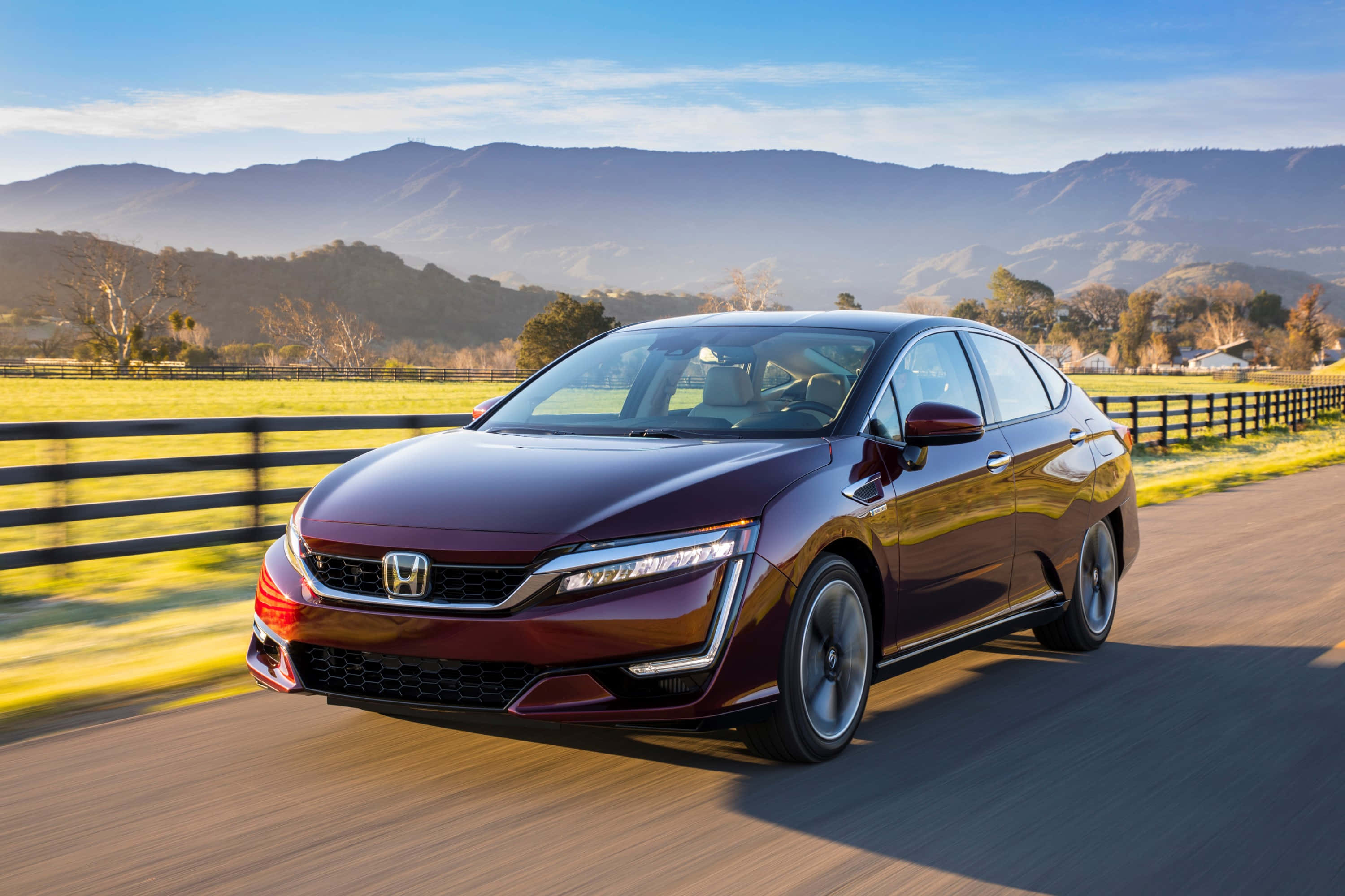 Honda Clarity 桌布 壁紙 — 一輛栗色本田房車在陽光明媚的鄉間道路上疾馳，沿途有起伏的丘陵和遠處的山脈，天空清澈湛藍。