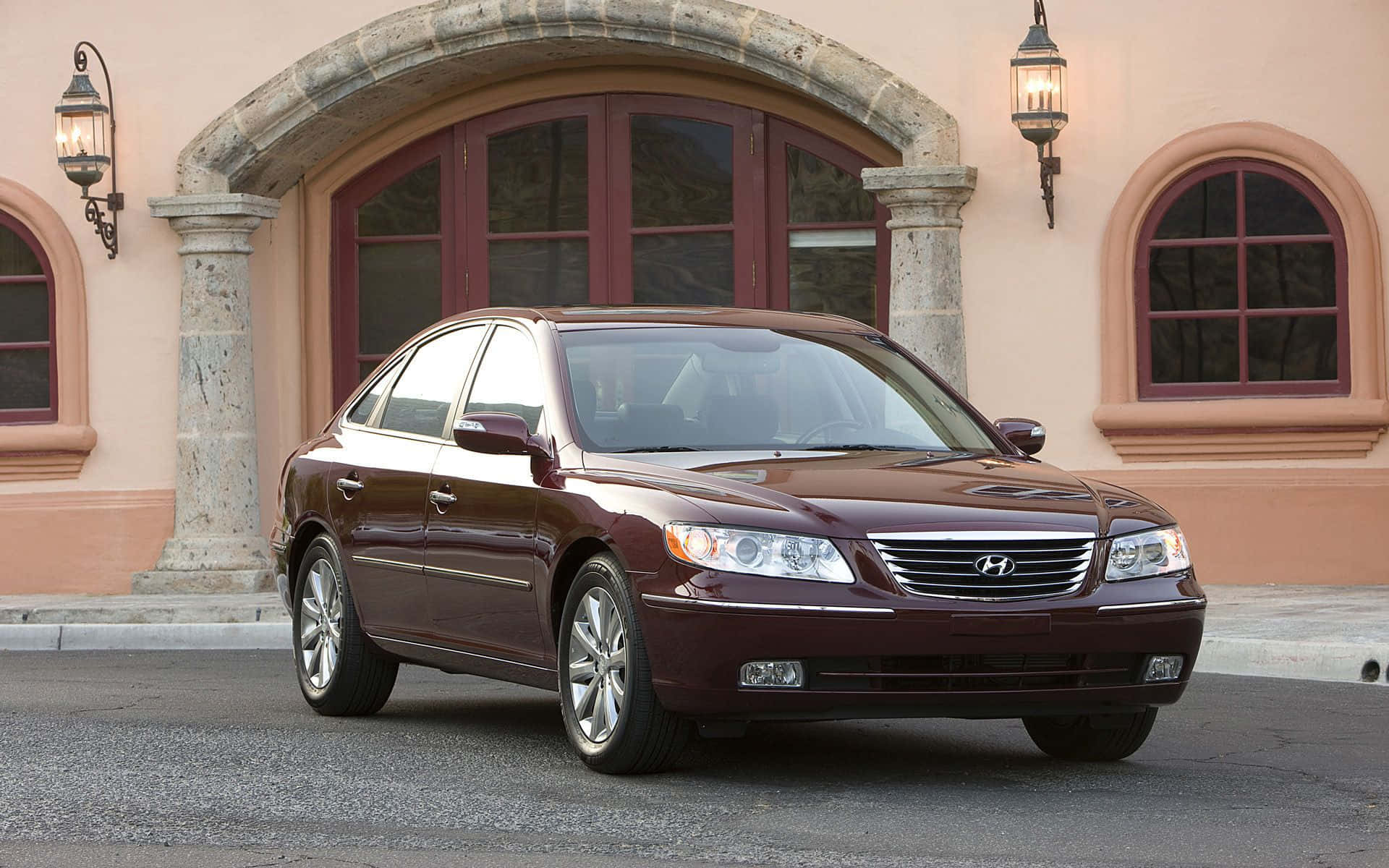 Hyundai Azera 桌布 壁紙 — 一輛栗色現代轎車在日光下閃閃發光，停在帶有拱形窗戶和石燈籠的灰泥建築前。