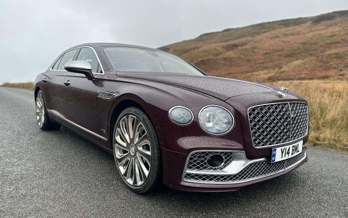 Bentley Flying Spur 桌布 壁紙 — 一輛栗色賓利飛馳在濕滑的道路上閃閃發光，背景是陰天下一片連綿的棕色山丘。