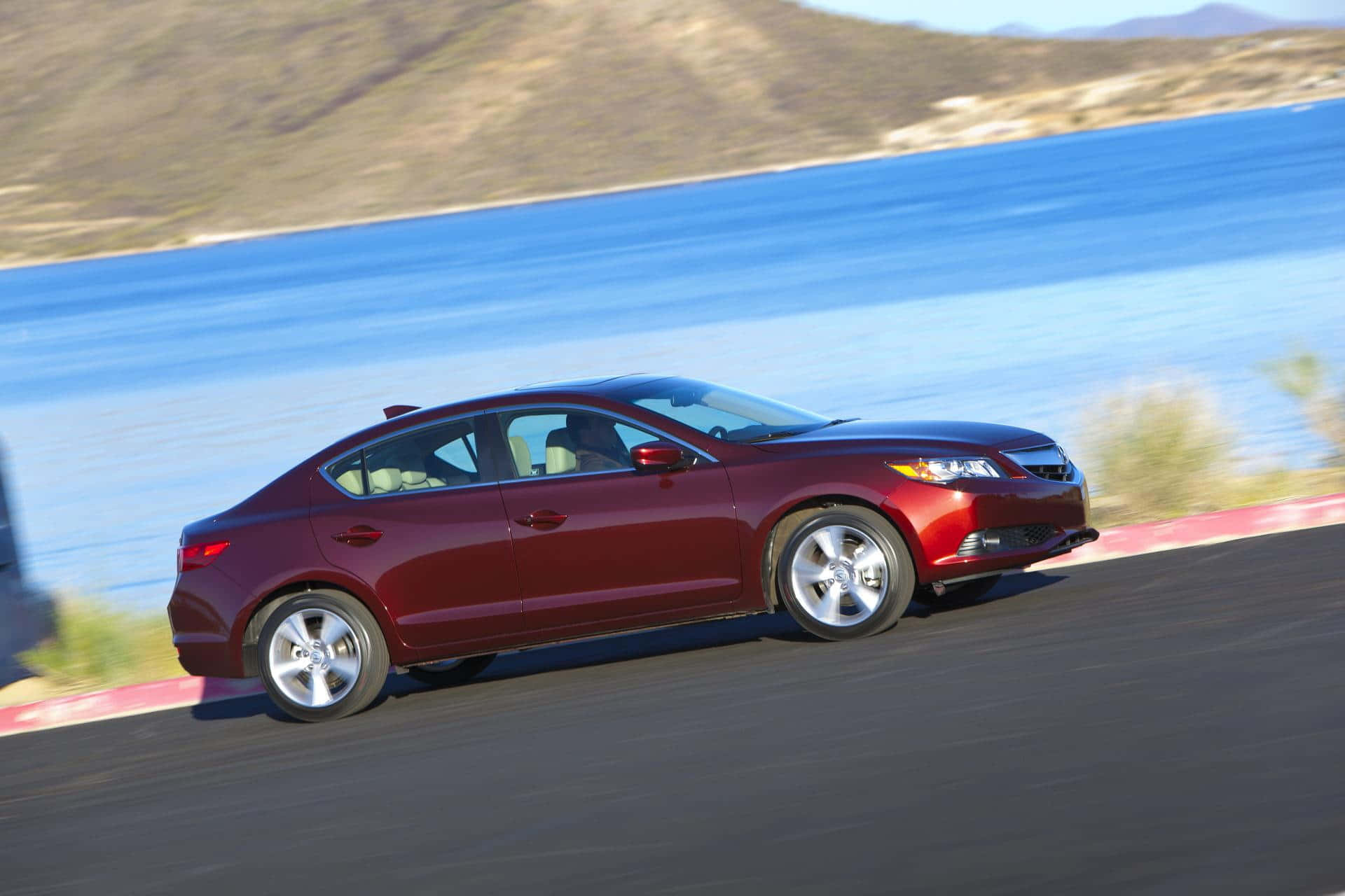 Acura Ilx 桌布 壁紙 — 一輛栗色轎車沿著海岸公路疾馳，背景是模糊的藍色海水和乾燥的山丘。