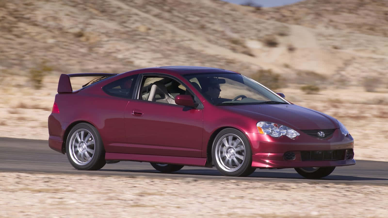 Acura Rsx 桌布 壁紙 — 一輛栗色運動轎跑車在明亮的日光下，沿著沙漠公路疾馳，背景是沙丘。