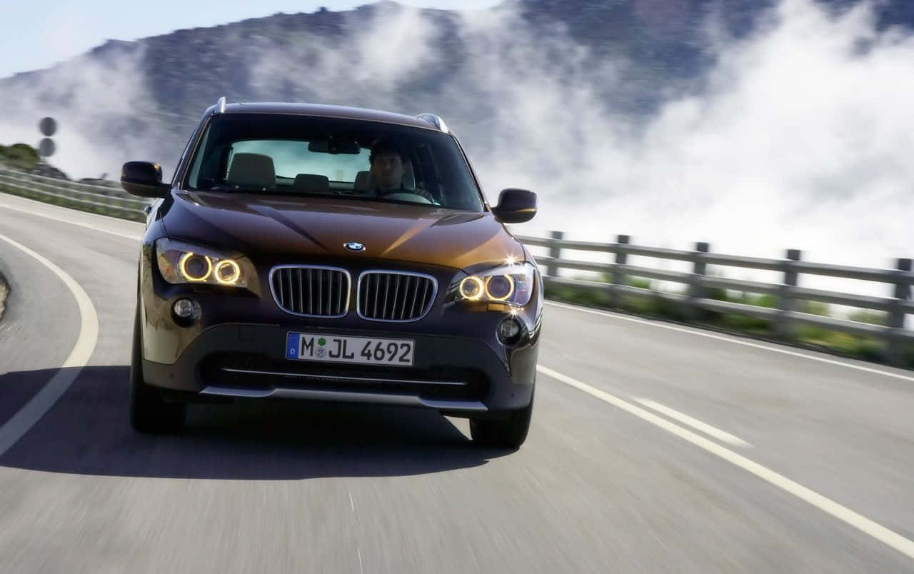 Bmw X1 桌布 壁紙 — 一輛棕色BMW X1 SUV在蜿蜒的山路上疾馳，山頂周圍有霧氣繚繞。