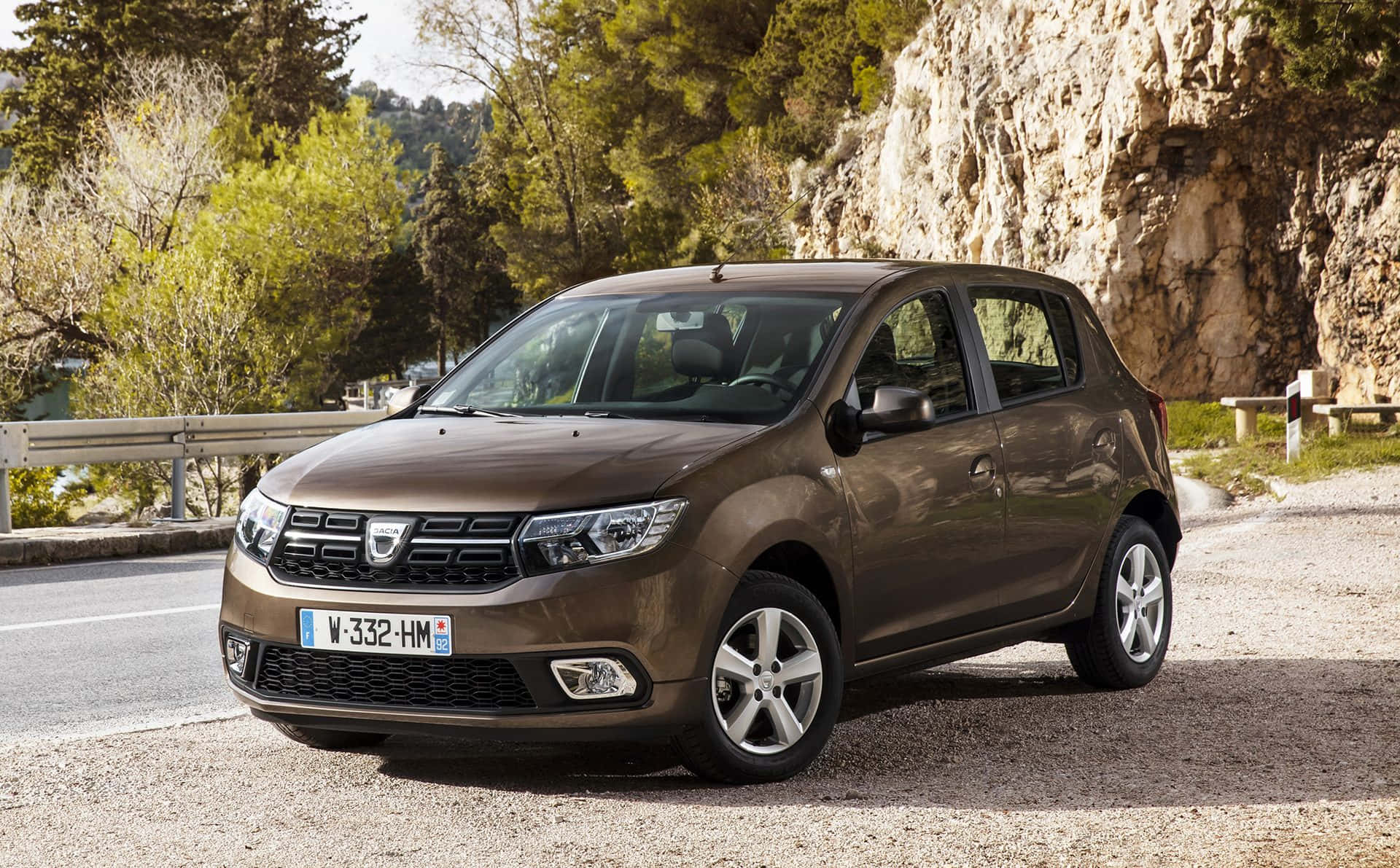 Dacia 桌布 壁紙 — 一輛棕色Dacia Sandero停泊在碎石路邊，旁邊是岩石懸崖和綠樹，在明亮的日光下。