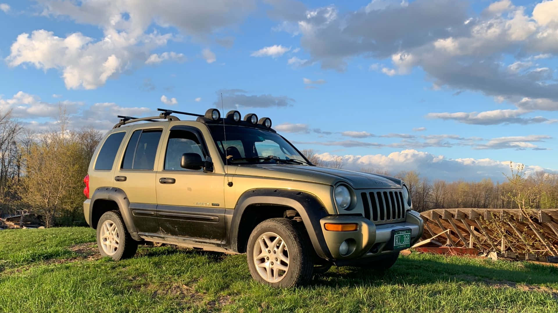 Jeep Liberty 桌布 壁紙 — 一輛棕褐色Jeep Liberty配備車頂行李架和聚光燈，停在草地上，天空部分多雲。