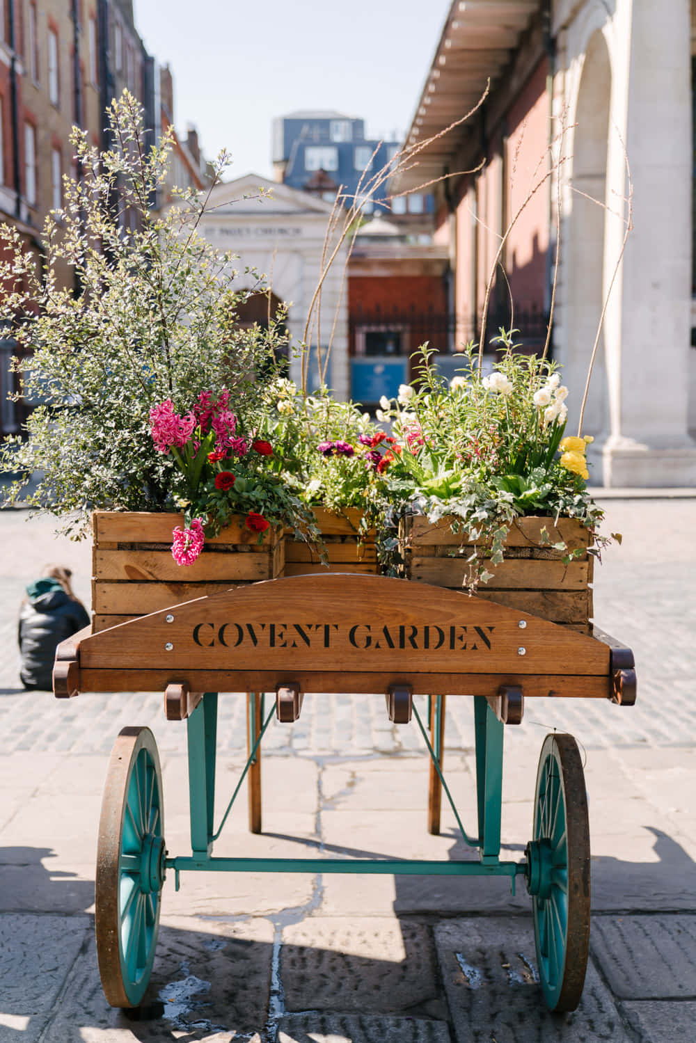 Flower Cart 桌布 壁紙 — 一輛標有Covent Garden的復古木製手推車，在陽光明媚的鵝卵石街道上裝滿了色彩繽紛的鮮花和綠植。