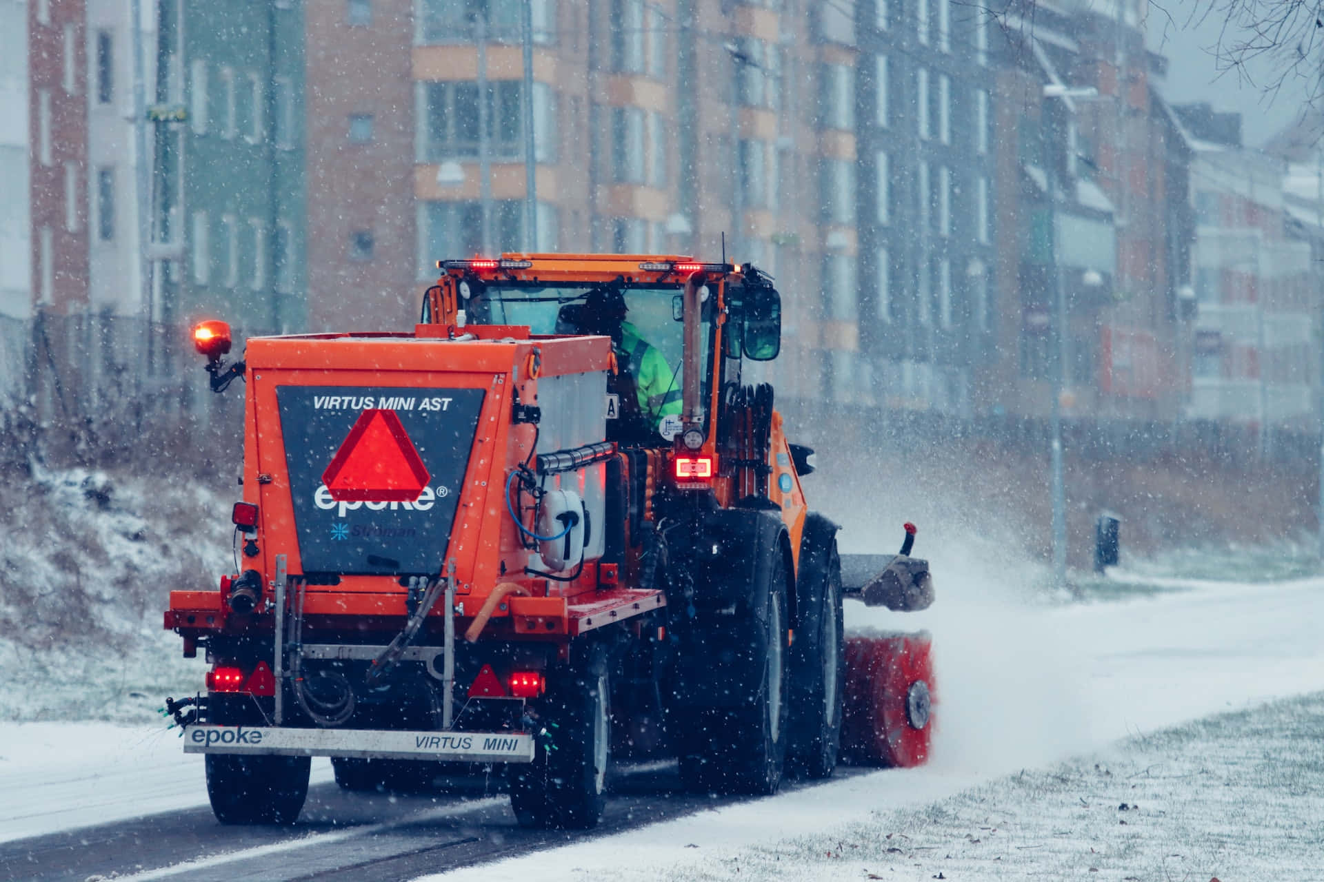 Snowplow 桌布 壁紙 — 一輛橙色剷雪車在積雪的城市街道上剷雪，雪花在模糊的公寓大樓背景下飄落。
