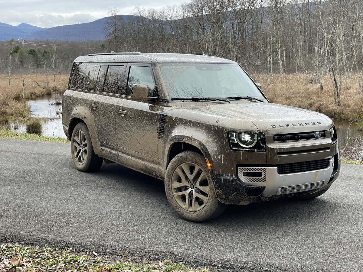 Land Rover Defender 桌布 壁紙 — 一輛沾滿泥濘的 Land Rover Defender 在濕滑的道路上行駛，穿越一片荒涼的冬季森林，遠方有山巒。