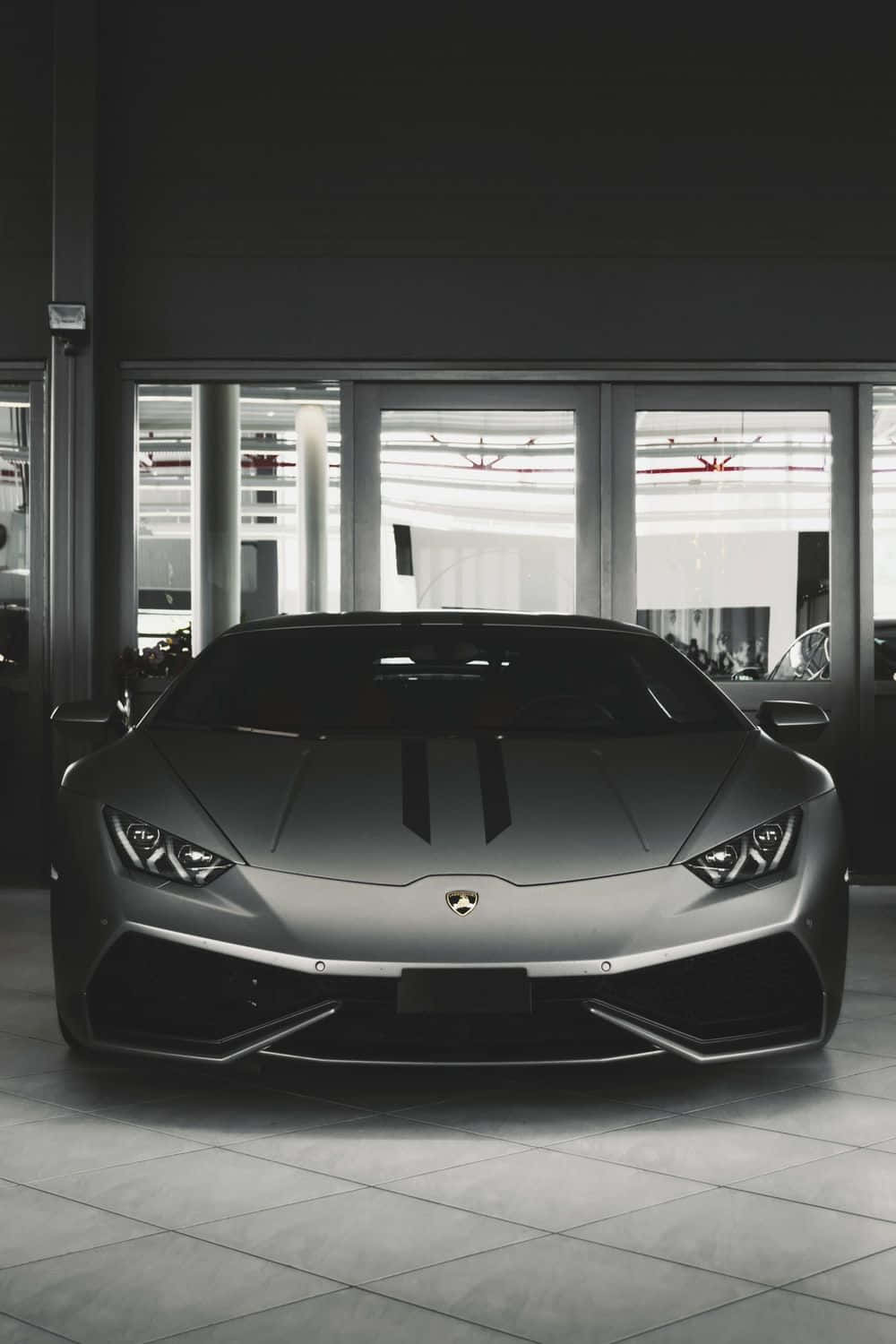 Black And White Car 桌布 壁紙 — 一輛流線型啞光灰色林寶堅尼跑車帶有黑色賽車條紋，位於現代簡約的展廳中央，地板鋪有瓷磚，窗戶寬大。
