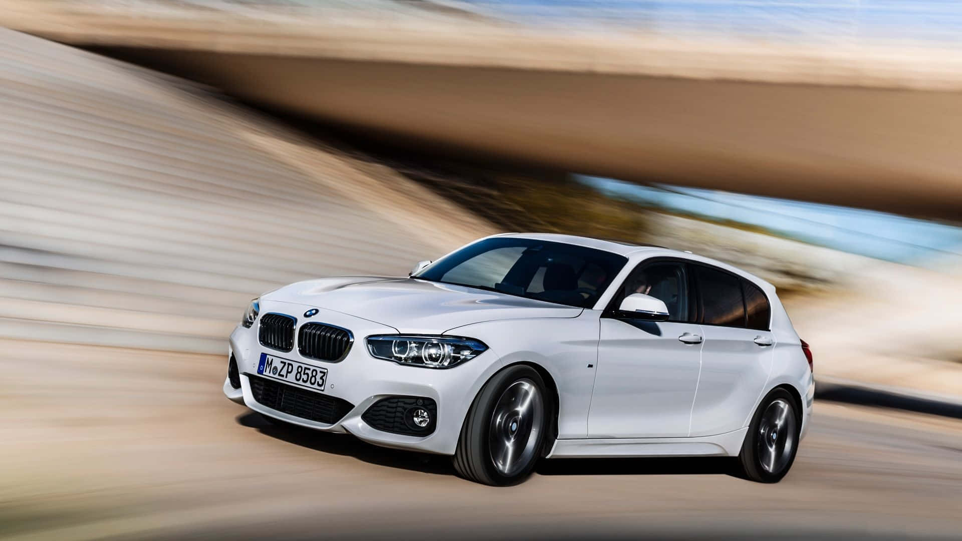 Bmw 1 Series 桌布 壁紙 — 一輛流線型白色BMW掀背車在陽光普照的道路上，於混凝土天橋下疾馳，背景帶有動態模糊效果。