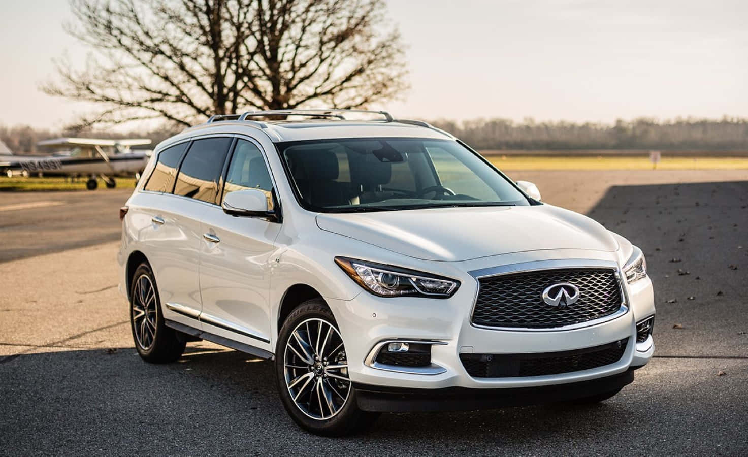 Infiniti Qx60 桌布 壁紙 — 一輛流線型白色Infiniti SUV停在瀝青路上，背景有小飛機和光禿禿的樹木，在柔和的金色光線下。