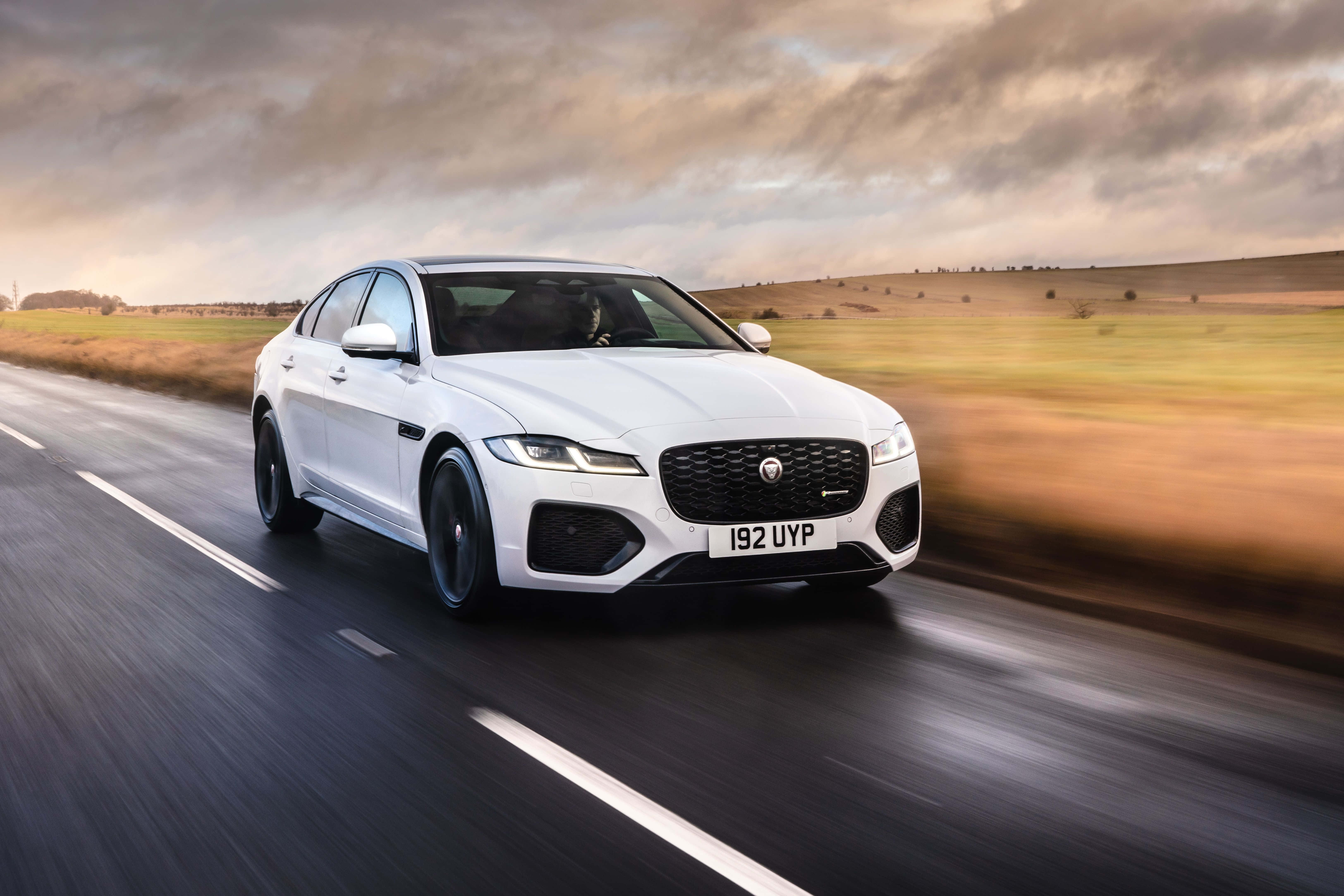 Jaguar Xf 桌布 壁紙 — 一輛流線型白色積架XF在鄉間道路上疾馳，背景是模糊的金色田野和動感的陰天。