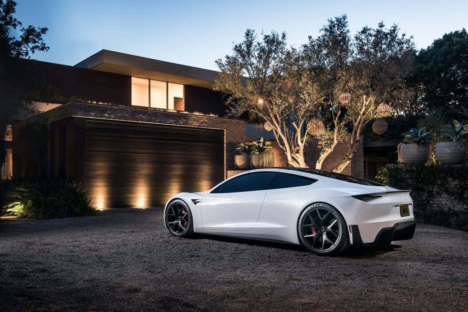 tesla 桌布 壁紙 — 一輛流線型白色跑車停泊在現代房屋前的碎石路上，黃昏時分，窗戶和樹木中透出溫暖的燈光。