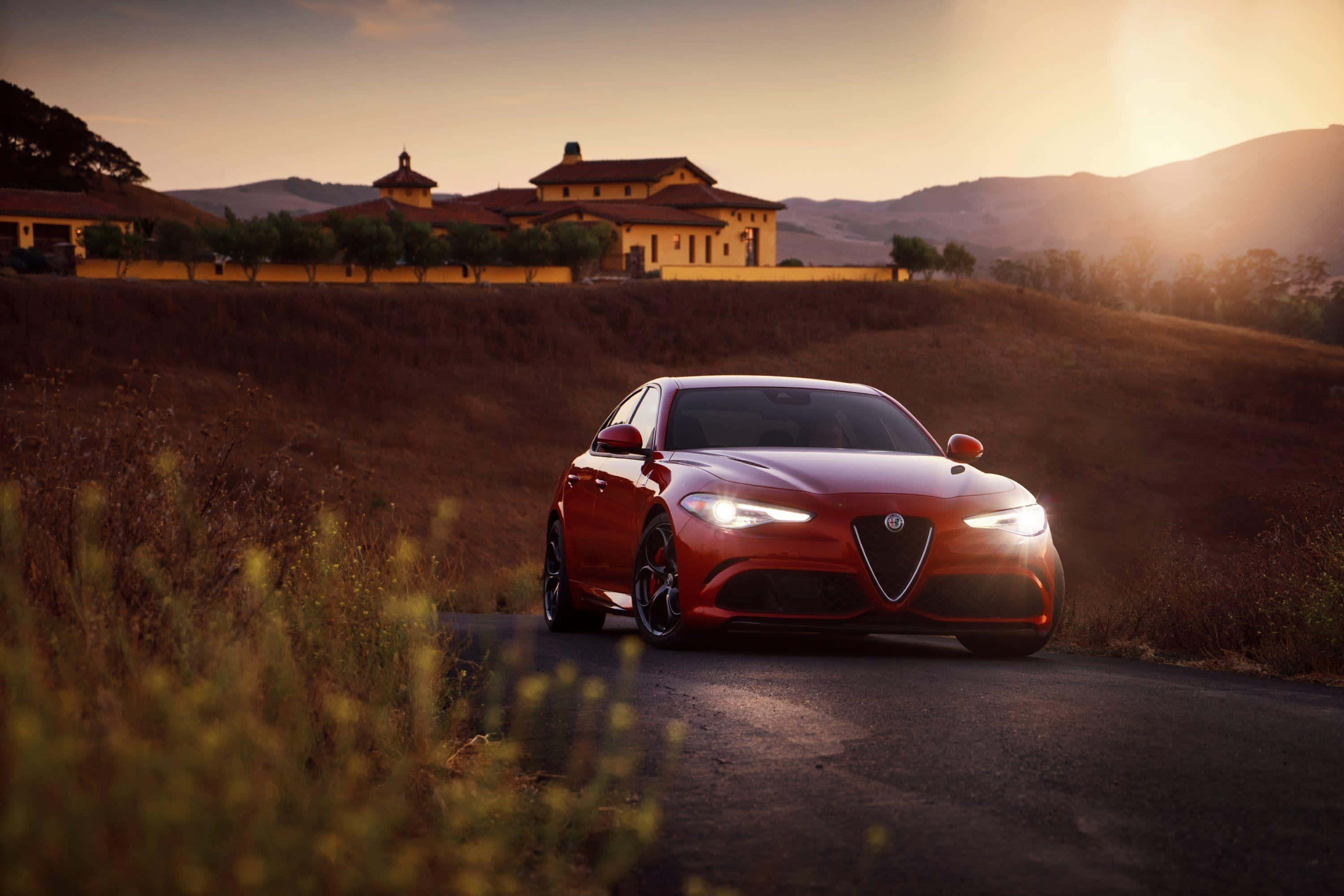 Alfa Romeo Giulia 桌布 壁紙 — 一輛流線型紅色跑車在日落時分沿著蜿蜒的道路行駛，金色光芒中映襯著質樸的別墅和起伏的山丘。