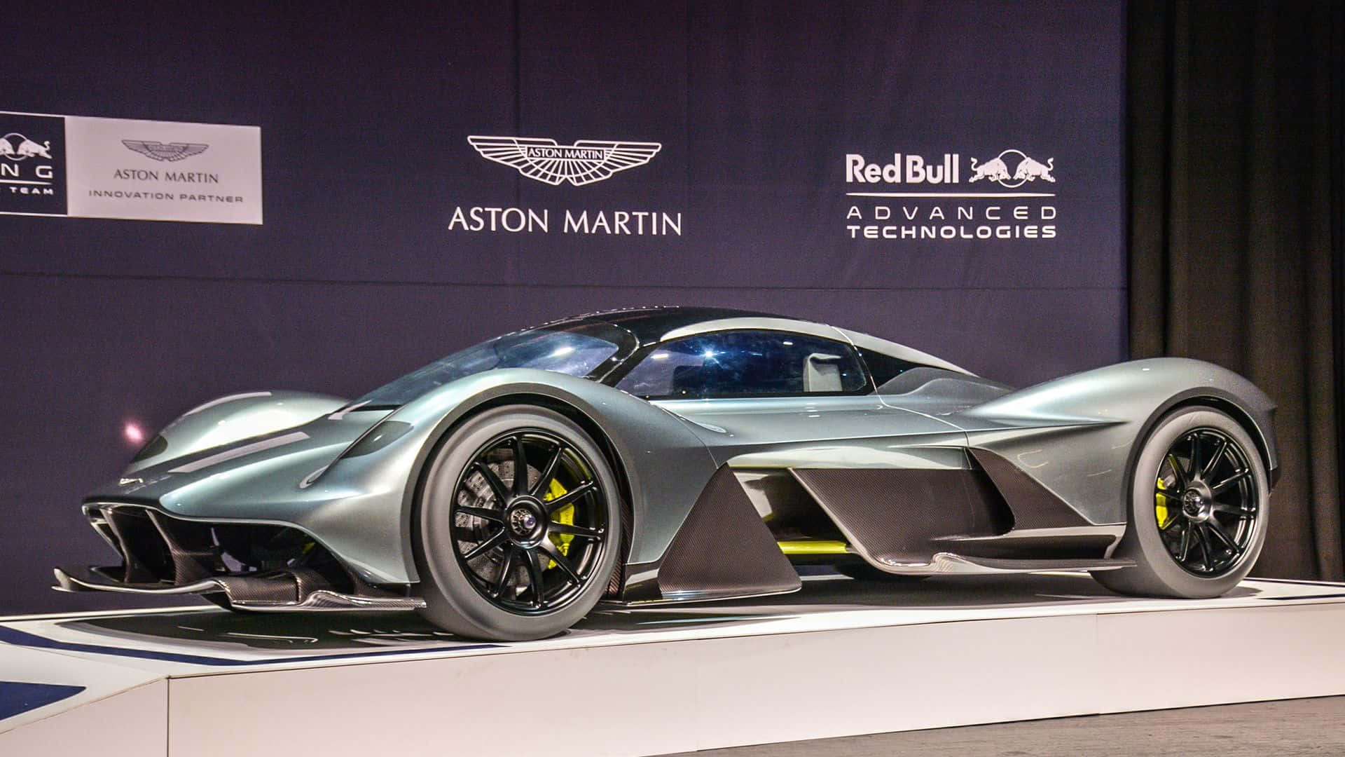 Aston Martin Valkyrie 桌布 壁紙 — 一輛流線型銀色Aston Martin hypercar配黃色制動卡鉗，在白色平台上展示，背景帶有品牌標誌。