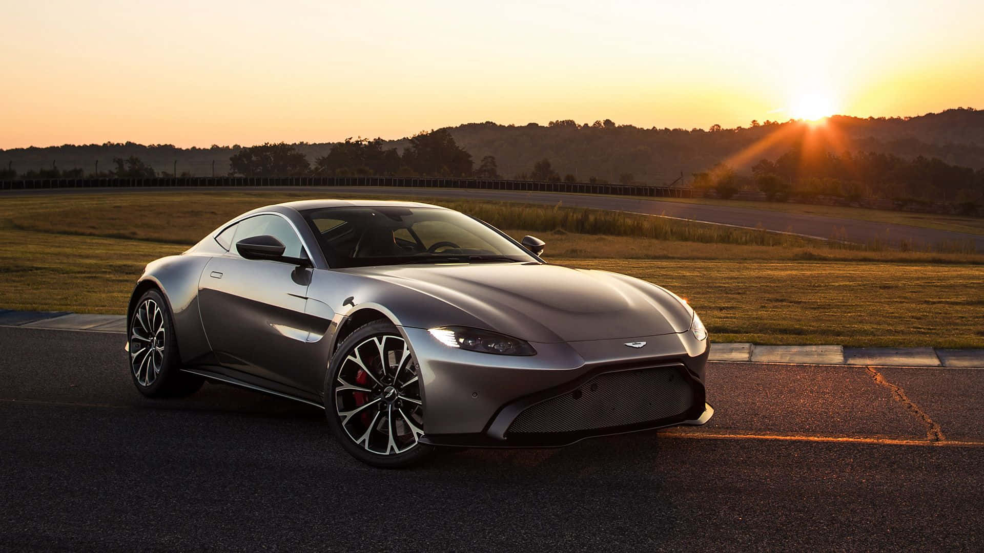 Aston Martin V12 Vantage 桌布 壁紙 — 一輛流線型銀色跑車在賽道上閃耀，夕陽在起伏的山丘後落下，灑下溫暖的金色光芒。