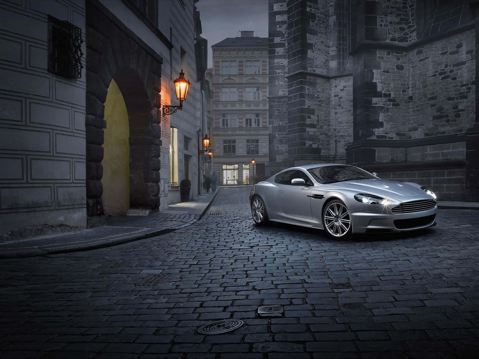 Aston Martin Db9 桌布 壁紙 — 一輛流線型銀色跑車在黃昏時分沿著鵝卵石小巷行駛，兩旁是歷史悠久的石砌建築和發光的燈籠。
