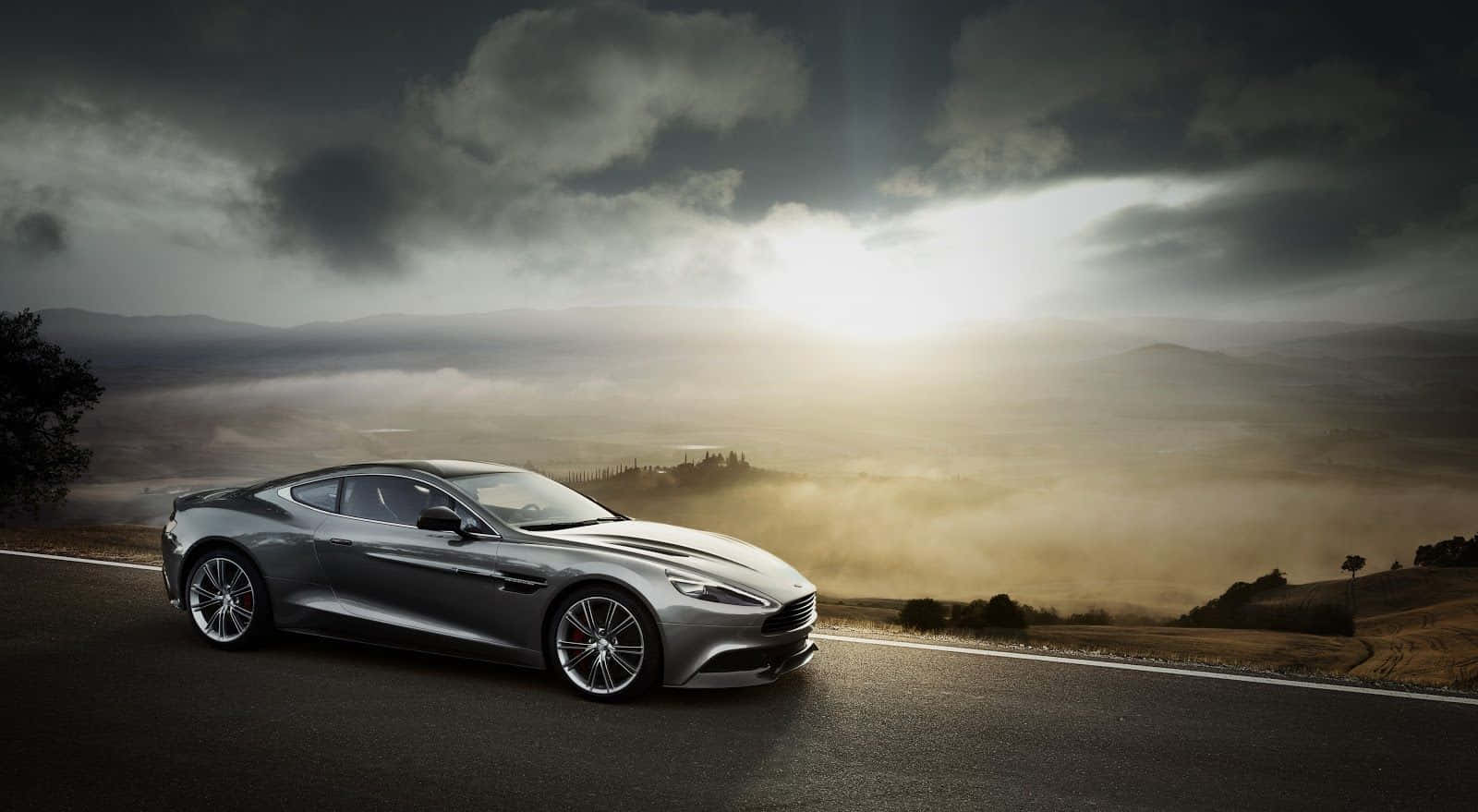 Aston Martin Rapide S 桌布 壁紙 — 一輛流線型銀色跑車沿著蜿蜒的山路行駛，金色陽光穿過暴風雲，照耀在霧氣瀰漫的山谷上。