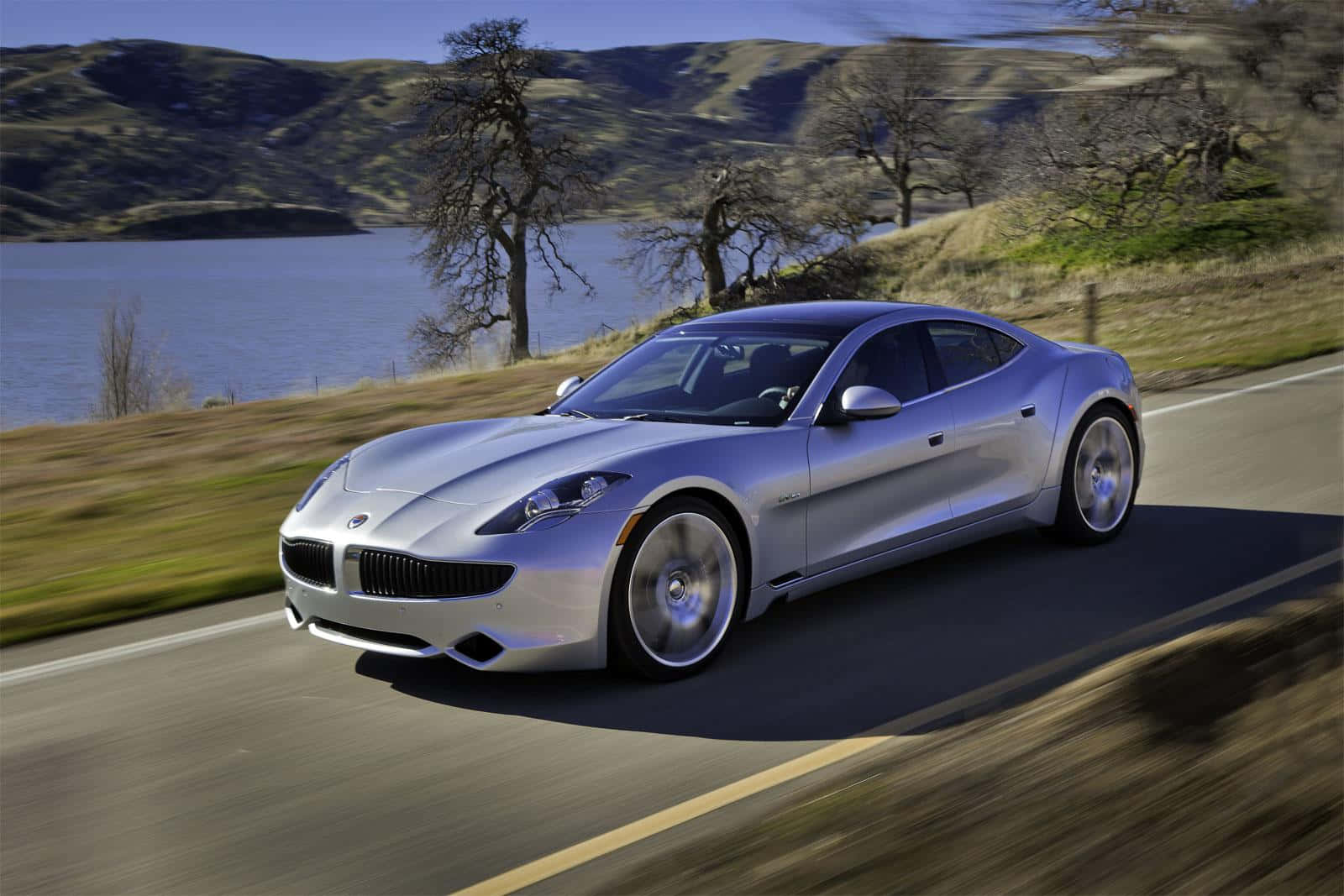 Fisker 桌布 壁紙 — 一輛流線型銀色跑車沿著蜿蜒的道路疾馳，旁邊是平靜的湖泊，背景是起伏的丘陵。