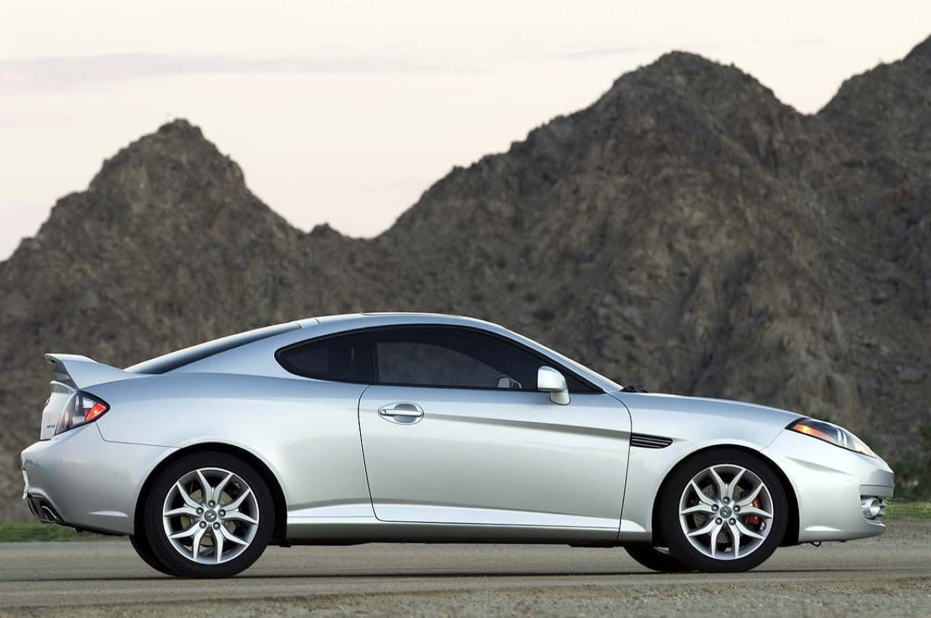 Hyundai Tiburon 桌布 壁紙 — 一輛流線型銀色運動轎跑車停在沙漠公路上，背景是鋸齒狀的岩石山脈，天空柔和暮色。