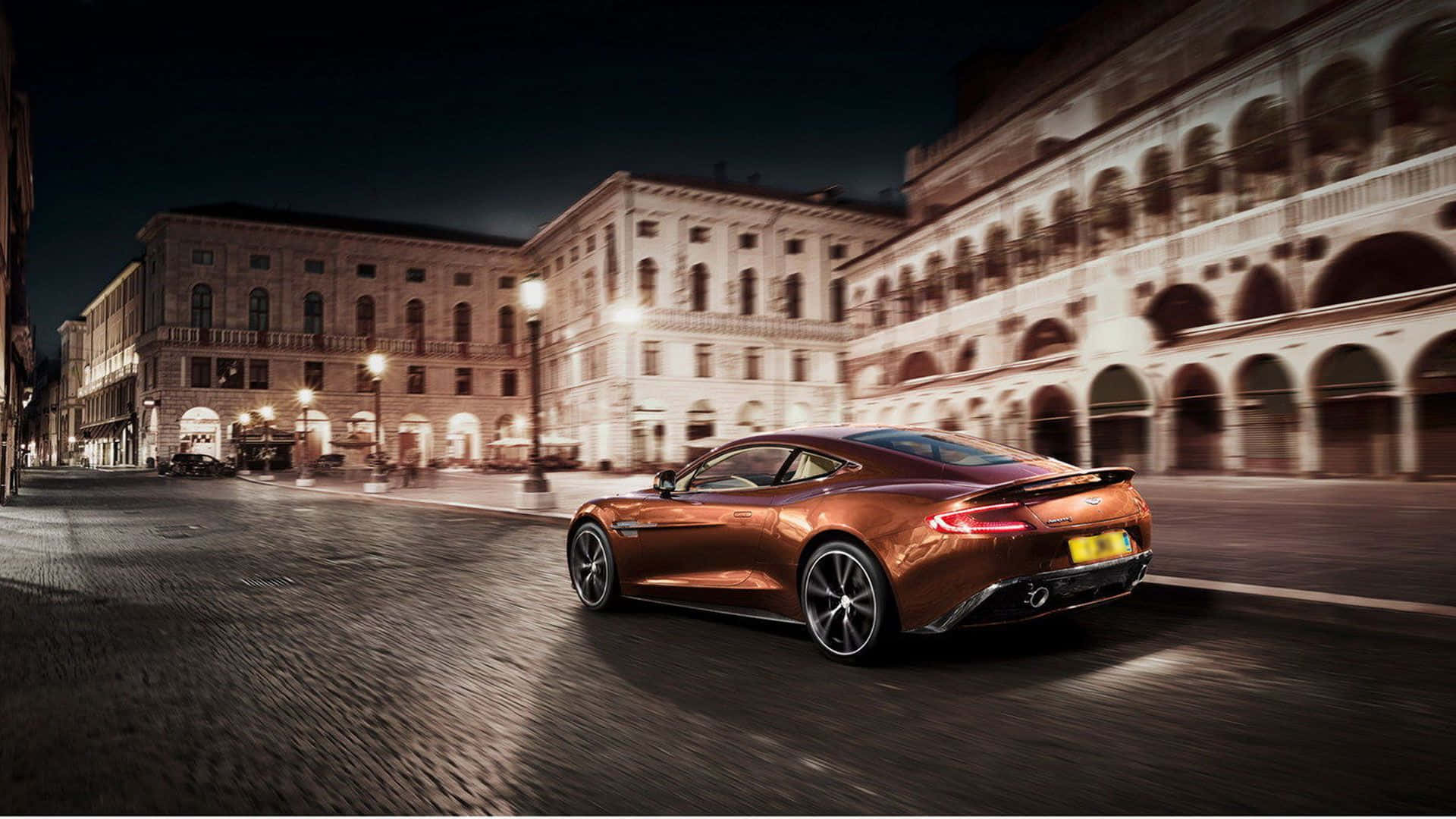 Aston Martin Vanquish 桌布 壁紙 — 一輛流線型青銅色跑車在夜間穿過歷史悠久的歐洲廣場，帶有動態模糊和溫暖的路燈光。