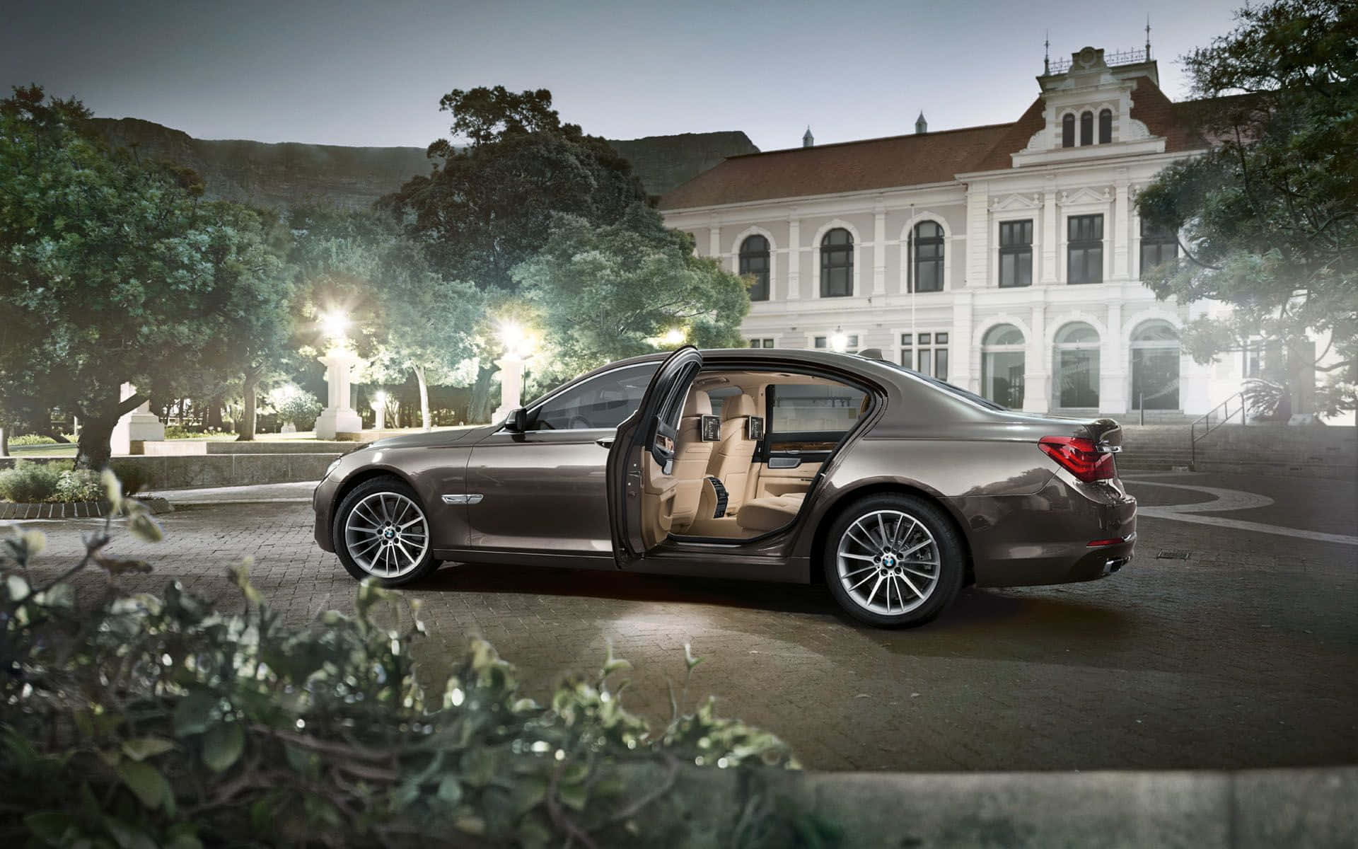 Bmw 7 Series 桌布 壁紙 — 一輛流線型青銅色轎車打開車門，露出棕色皮革內飾，黃昏時停在一棟有樹木和發光路燈的白色宏偉建築前。