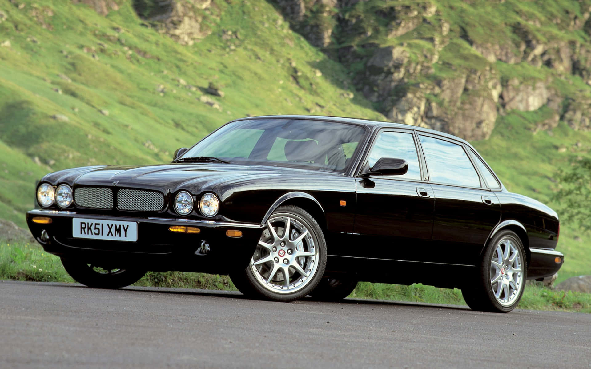 Jaguar Xjr 桌布 壁紙 — 一輛流線型黑色積架XJ房車沿著蜿蜒的山路行駛，背景是鬱鬱蔥蔥的綠色山丘和岩石峭壁。