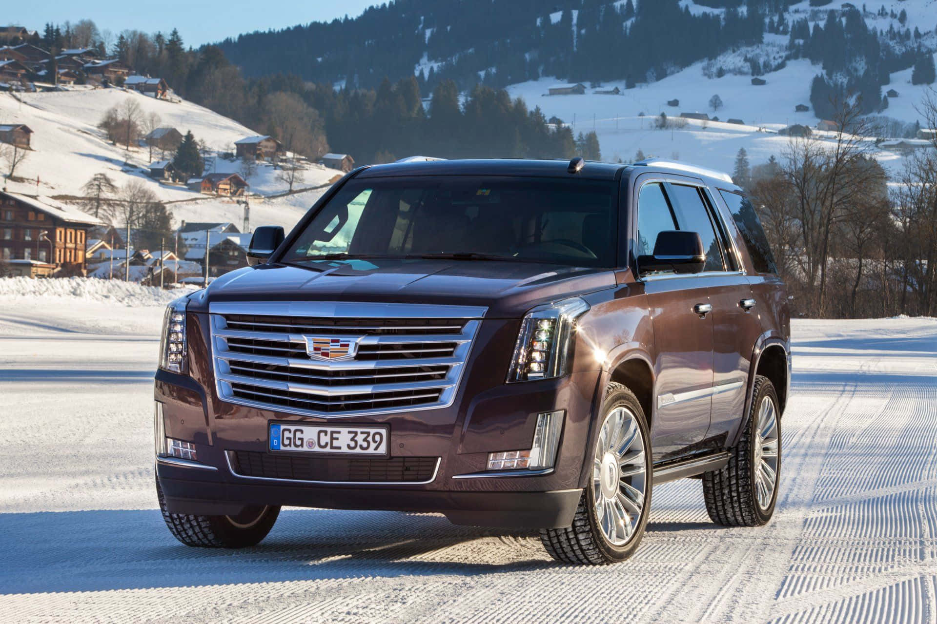 Cadillac Escalade 桌布 壁紙 — 一輛深啡色嘅Cadillac SUV喺一條積雪嘅路上行駛，穿過一個風景如畫嘅阿爾卑斯山村，背景係被雪覆蓋嘅山脈同木屋。