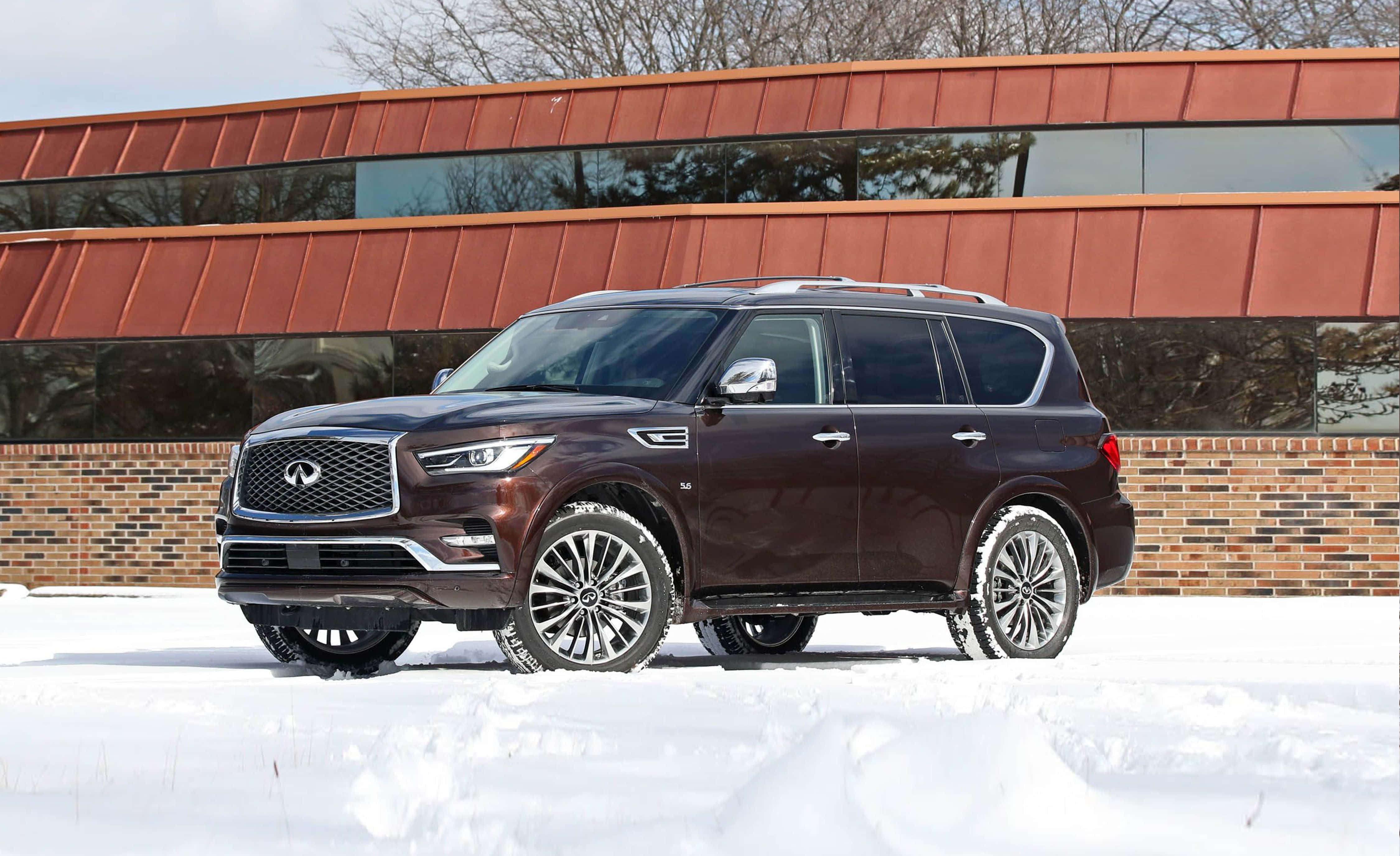 Infiniti Qx80 桌布 壁紙 — 一輛深棕色的 Infiniti QX80 SUV 停在新雪中，旁邊是一座現代建築，擁有銅色屋頂和玻璃窗。