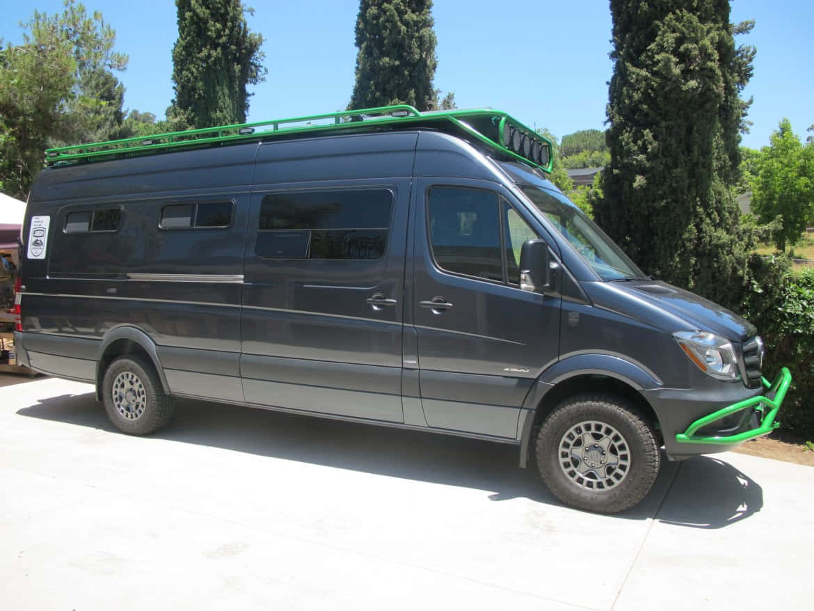 Mercedes Benz Sprinter 桌布 壁紙 — 一輛深灰色 Mercedes-Benz Sprinter 客貨車，帶有亮綠色車頂行李架和前保險槓，停泊在混凝土車道上，周圍是高聳的柏樹，天空清澈湛藍。