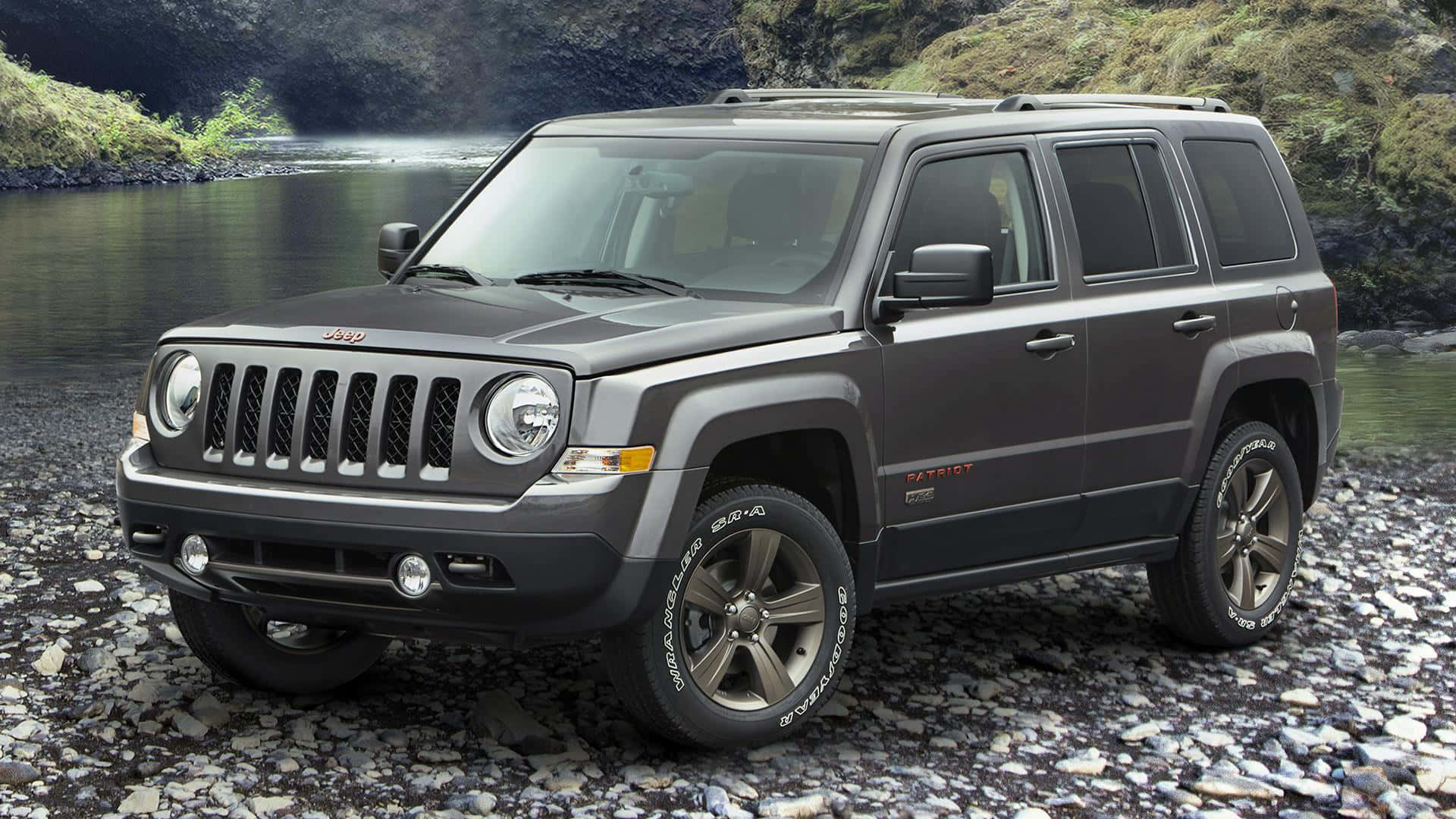 Jeep Patriot 桌布 壁紙 — 一輛深灰色Jeep Patriot SUV停泊在岩石河岸上，背景有綠色山丘和寧靜的水面。