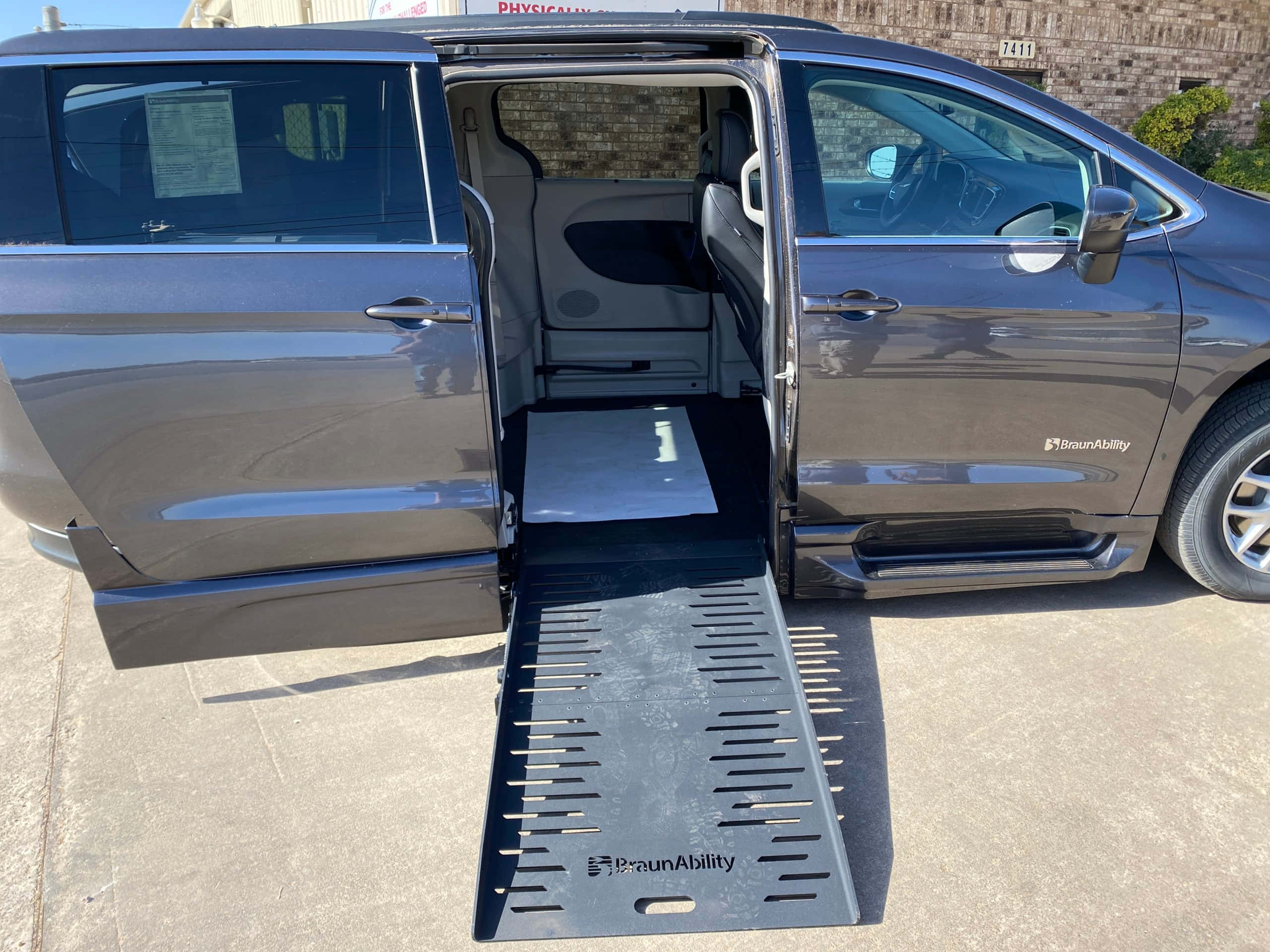 Chrysler Voyager 桌布 壁紙 — 一輛深灰色的BraunAbility客貨車，側門打開，金屬斜坡延伸到混凝土表面上，在明亮的陽光下。