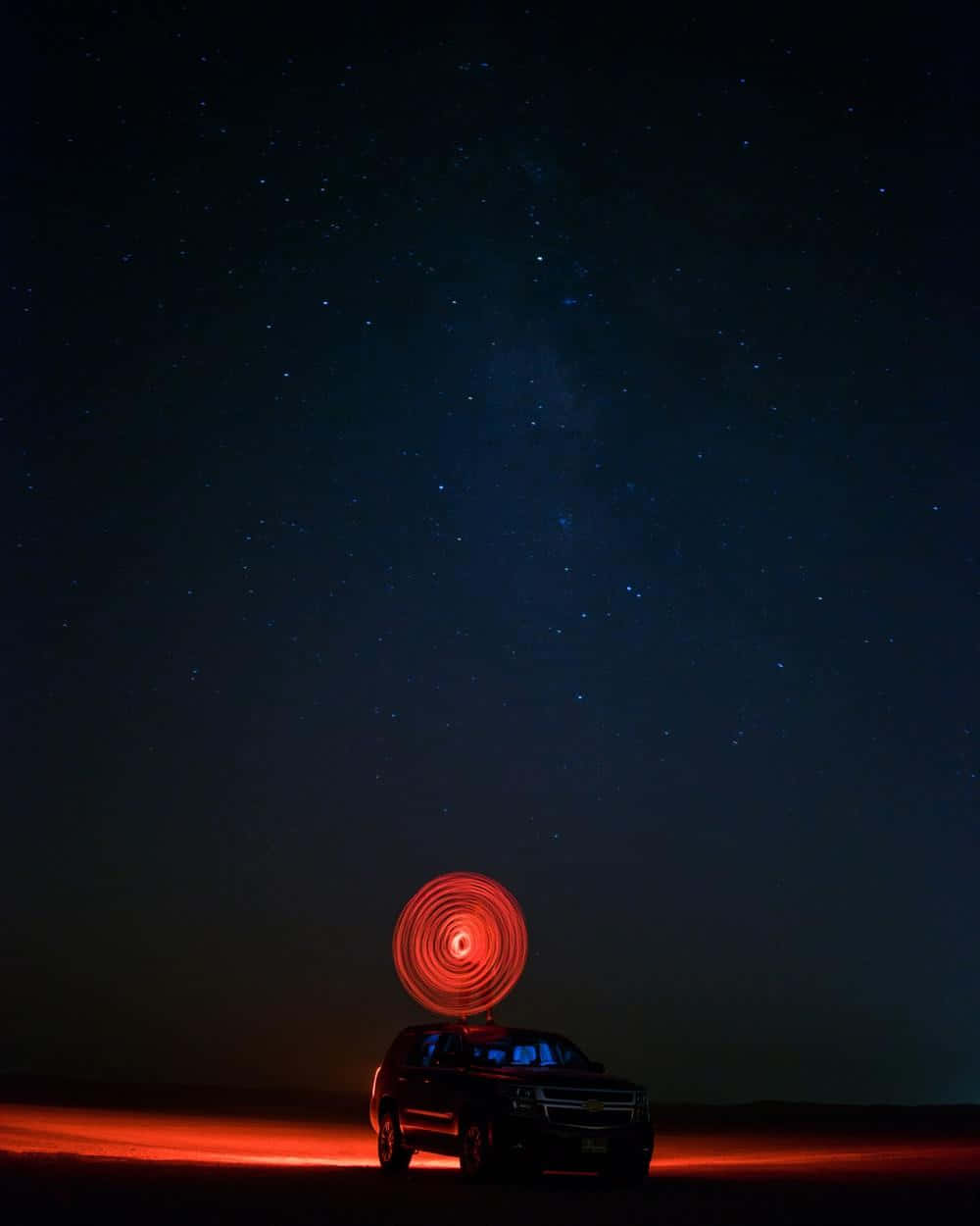深色手機 桌布 壁紙 — 一輛深色SUV停泊在繁星點點的夜空下，上方有發光的紅色螺旋光跡，地面有橙色照明。