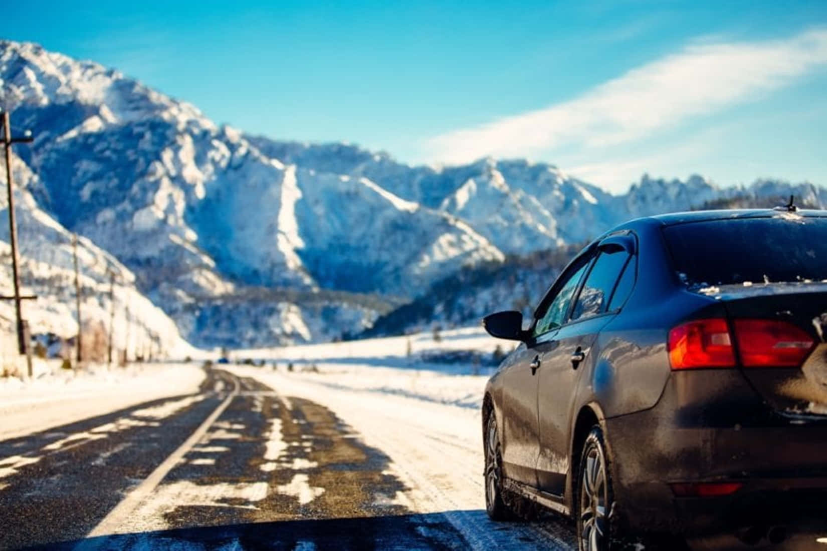 Winter Driving 桌布 壁紙 — 一輛深色轎車停泊在積雪的路邊，白雪覆蓋的山巒在清澈的藍天下聳立。