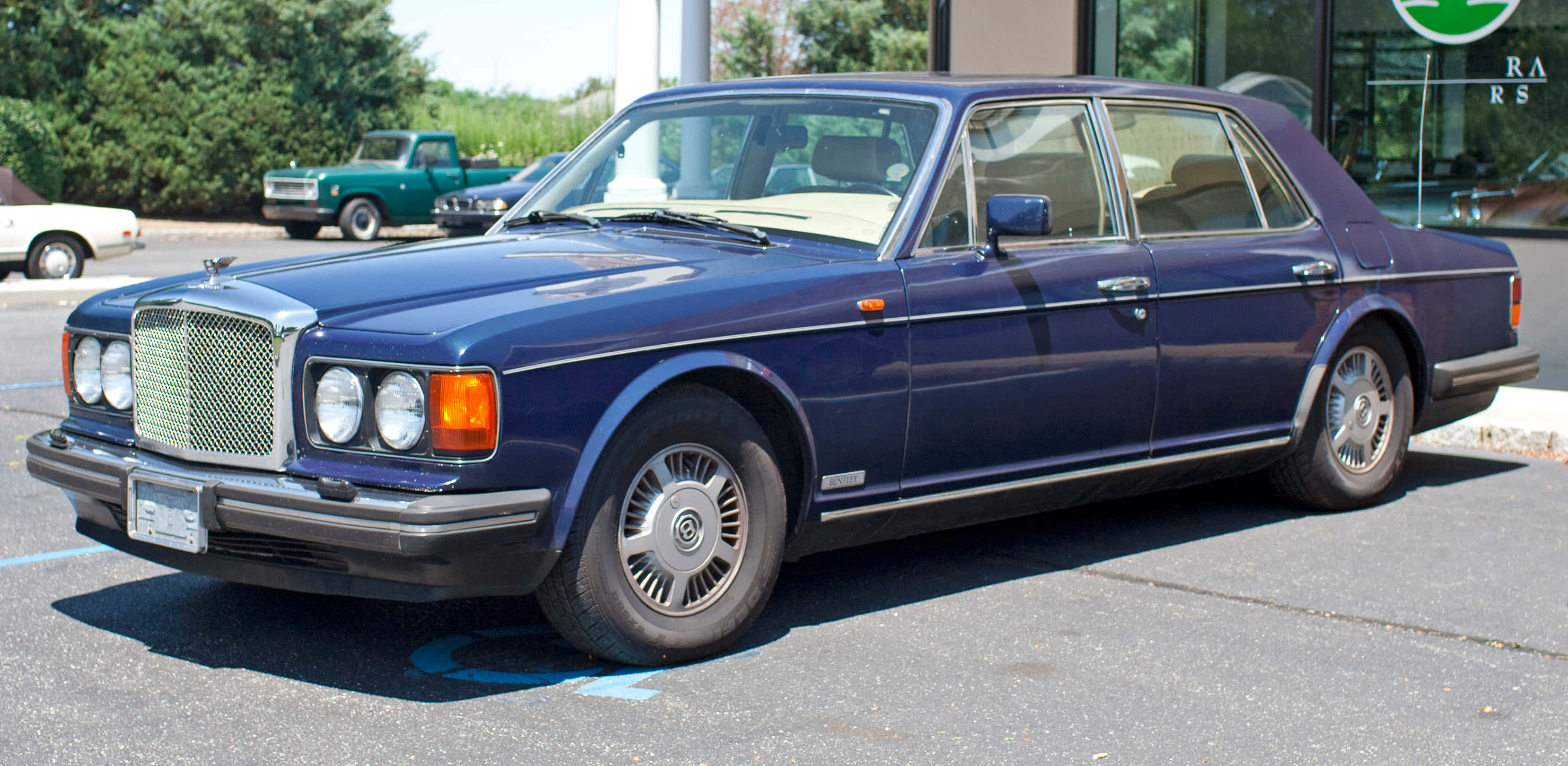 Bentley Eight 桌布 壁紙 — 一輛深藍色賓利房車在明亮陽光下閃閃發光，停放在一個附近有其他復古汽車的停車場。