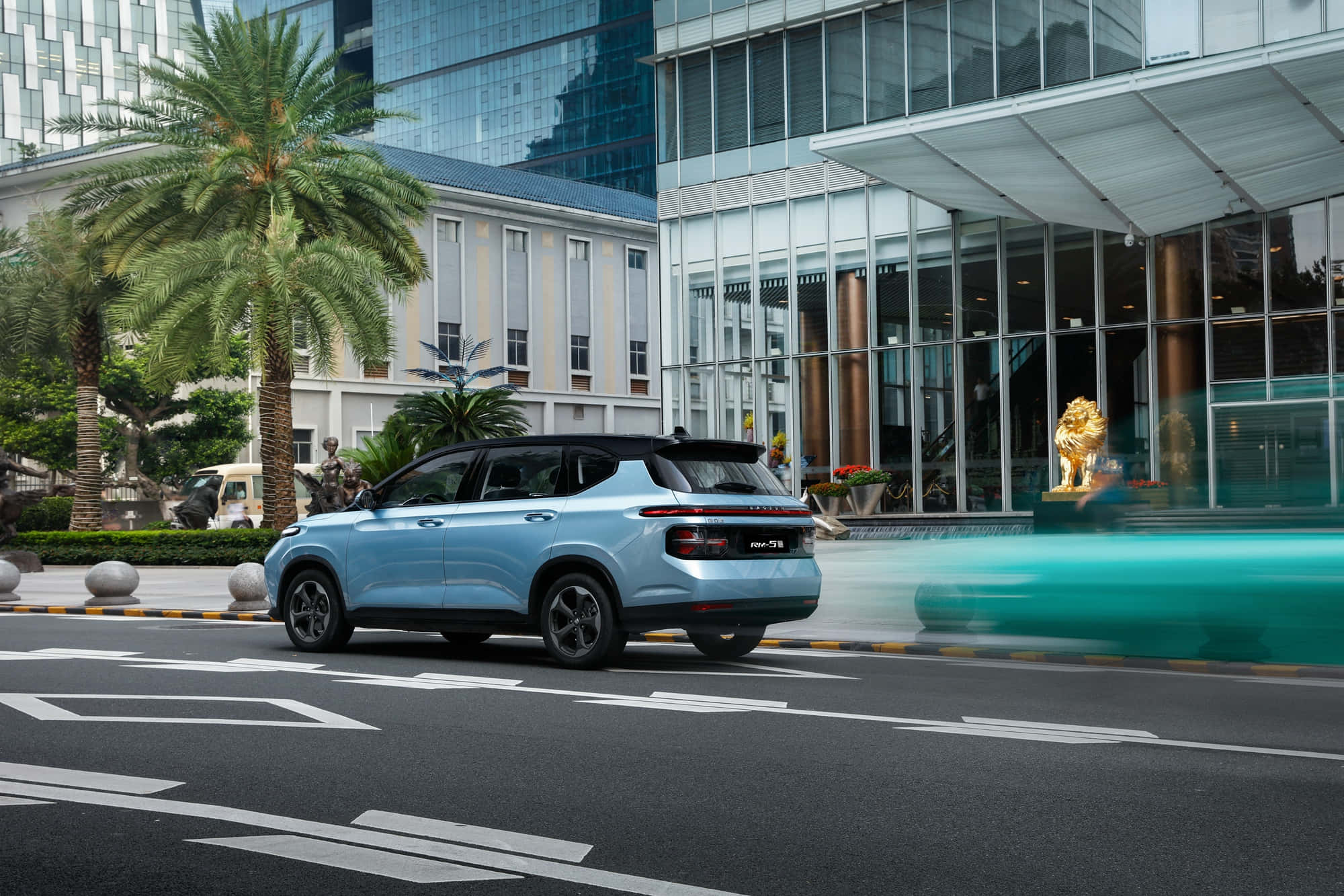 Baojun 桌布 壁紙 — 一輛淺藍色SUV駛過一棟現代玻璃建築，建築旁有棕櫚樹和一尊金色獅子雕像。