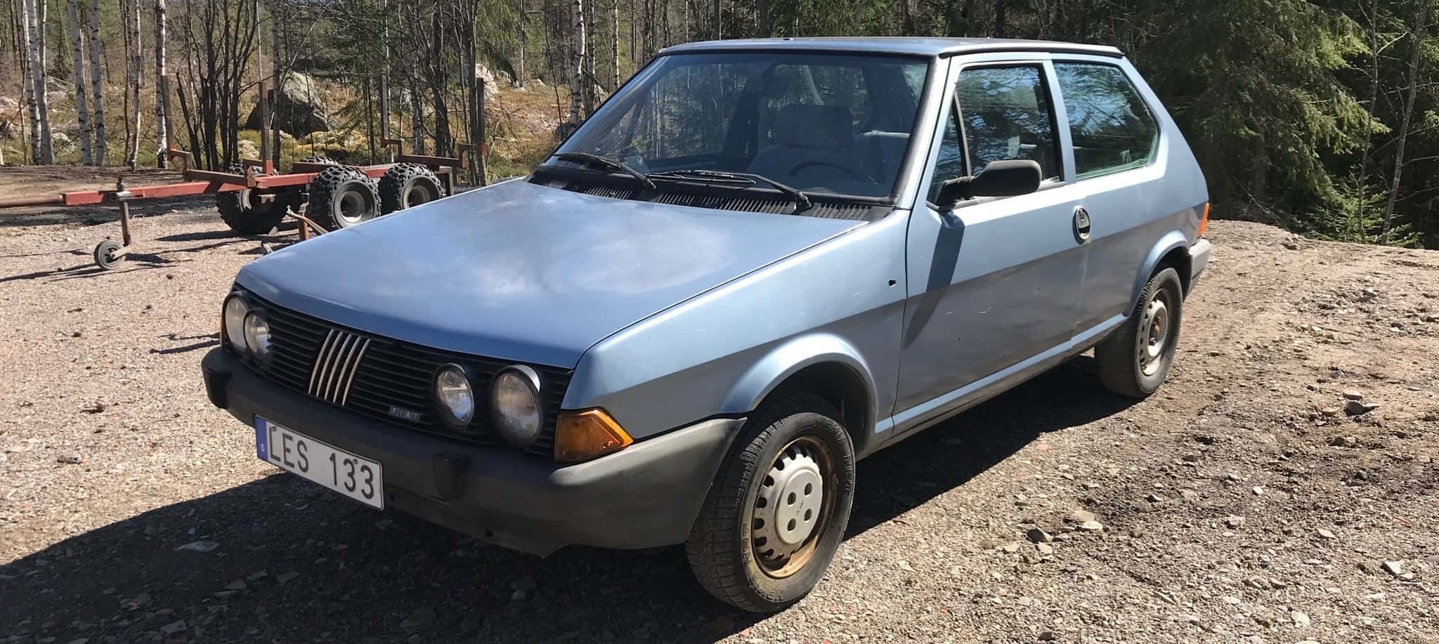 Fiat Ritmo 桌布 壁紙 — 一輛淺藍色復古菲亞特Uno停泊在碎石地上，四周是高聳的松樹，在明亮的日光下。