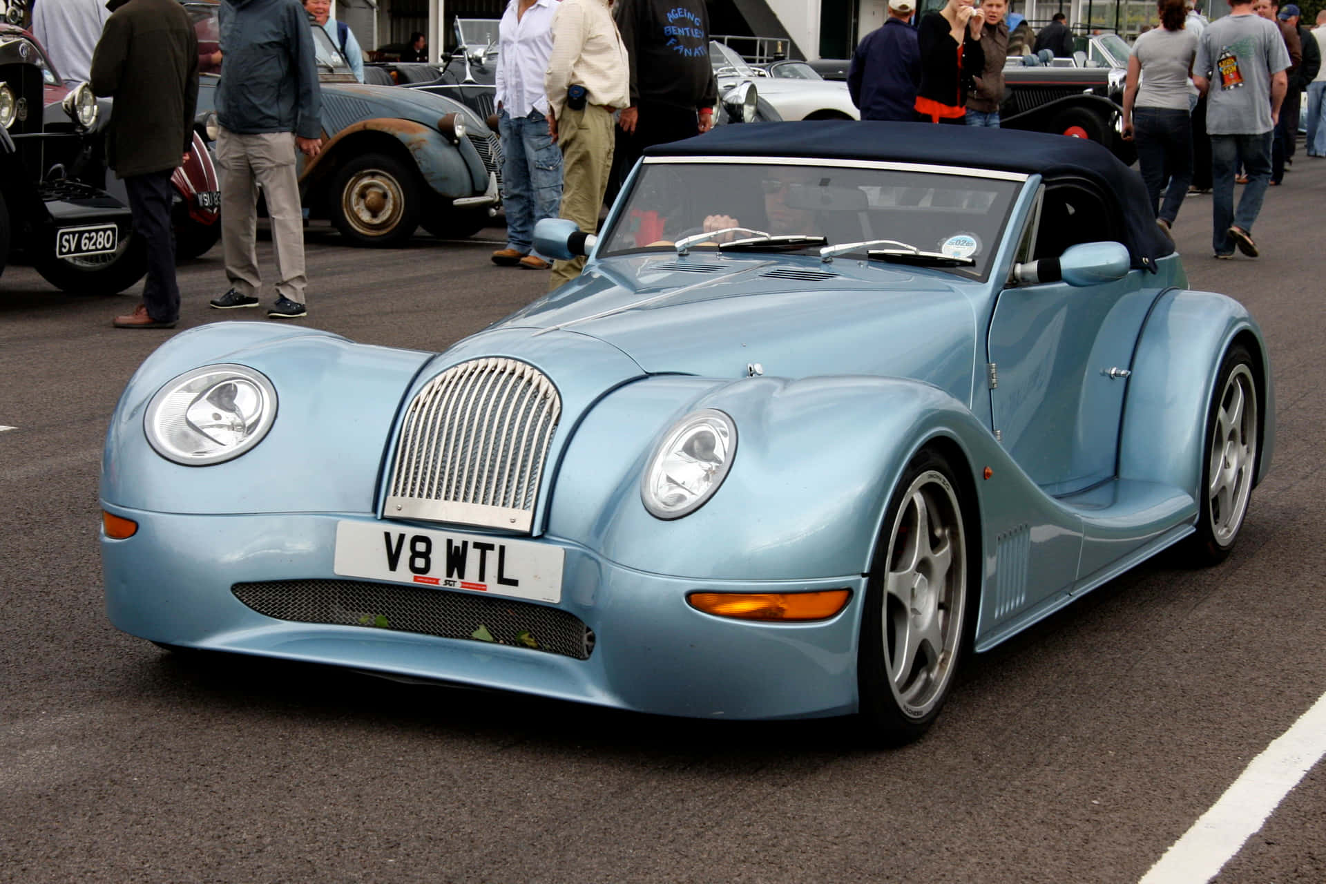 Morgan 桌布 壁紙 — 一輛淺藍色復古風格黑色軟頂開篷跑車停泊在車展上，周圍有圍觀者。