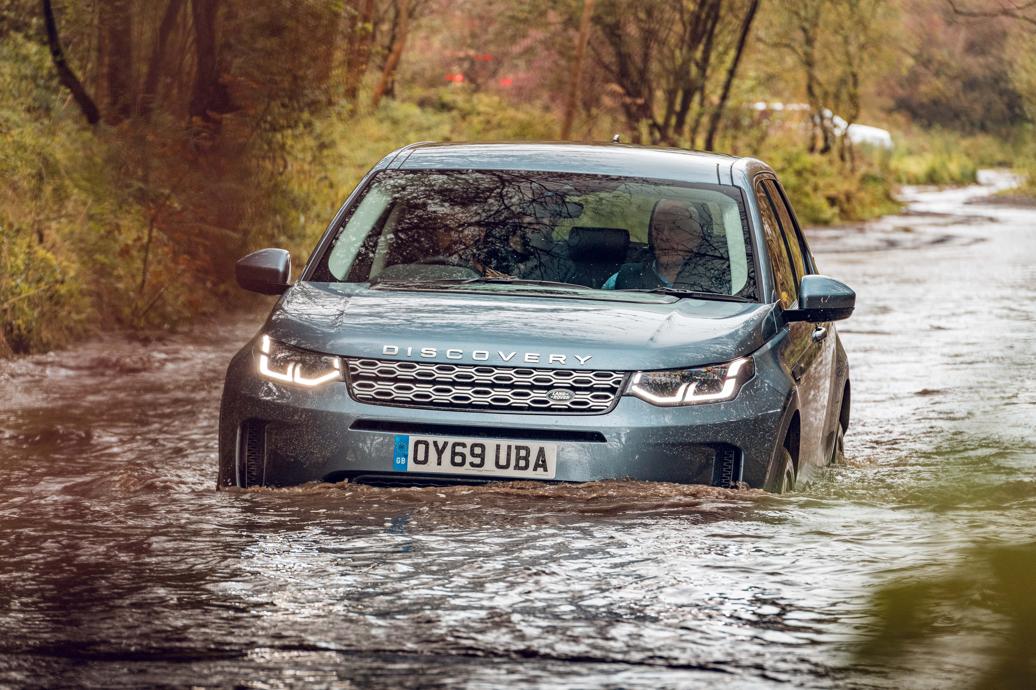 Land Rover Discovery 桌布 壁紙 — 一輛灰色 Land Rover Discovery 在泥濘的森林水坑中行駛，車頭燈亮起，潮濕的道路兩旁樹木林立。