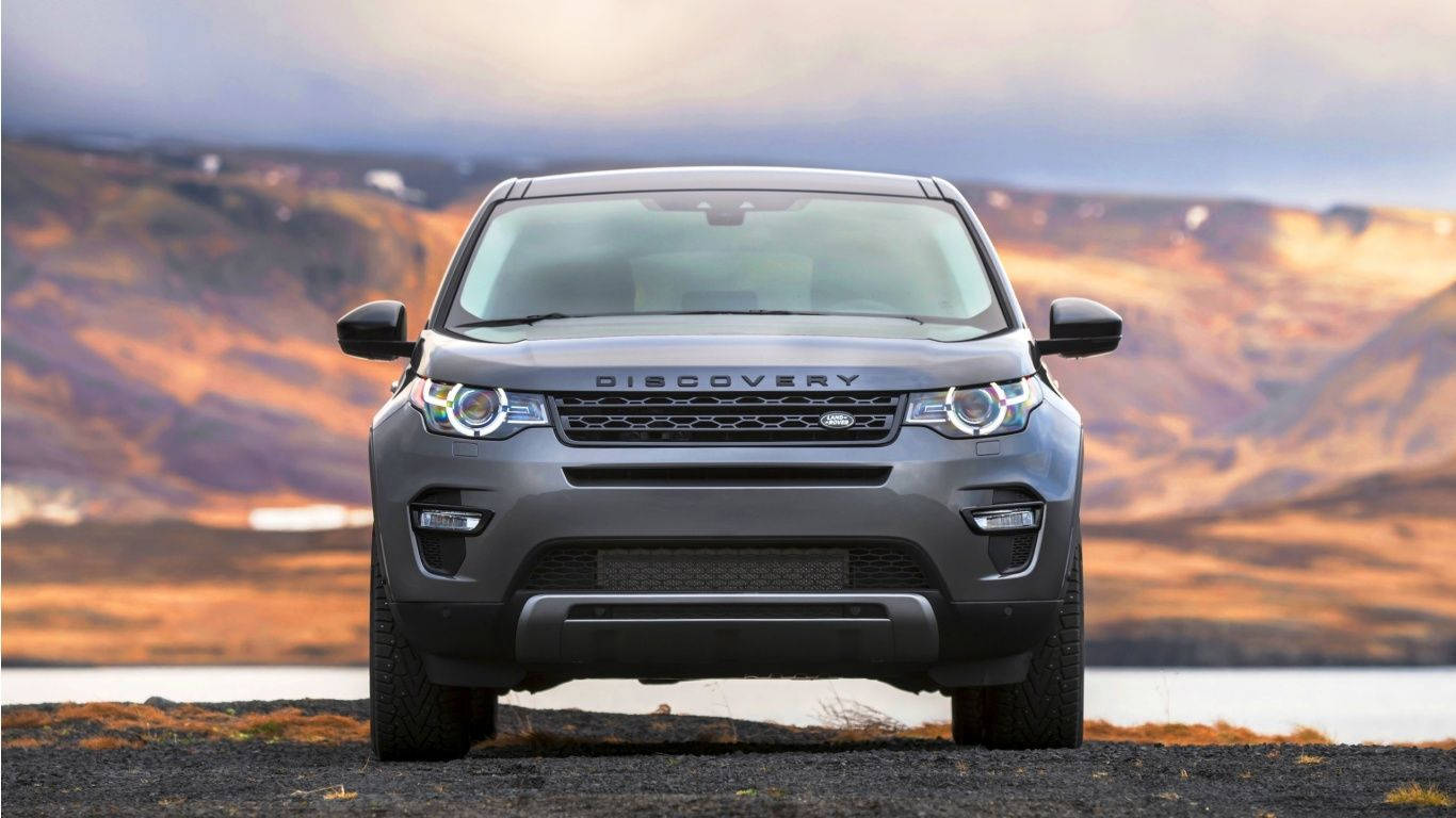 沙漠 桌布 壁紙 — 一輛灰色 Land Rover Discovery SUV 在碎石灘上向前行駛，背景是金色山丘和寧靜的湖泊，天空呈現柔和的暮色。